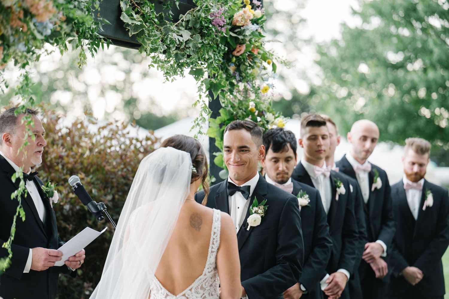 A romantic outdoor wedding ceremony under a floral arch with a bridal party in classic black suits.