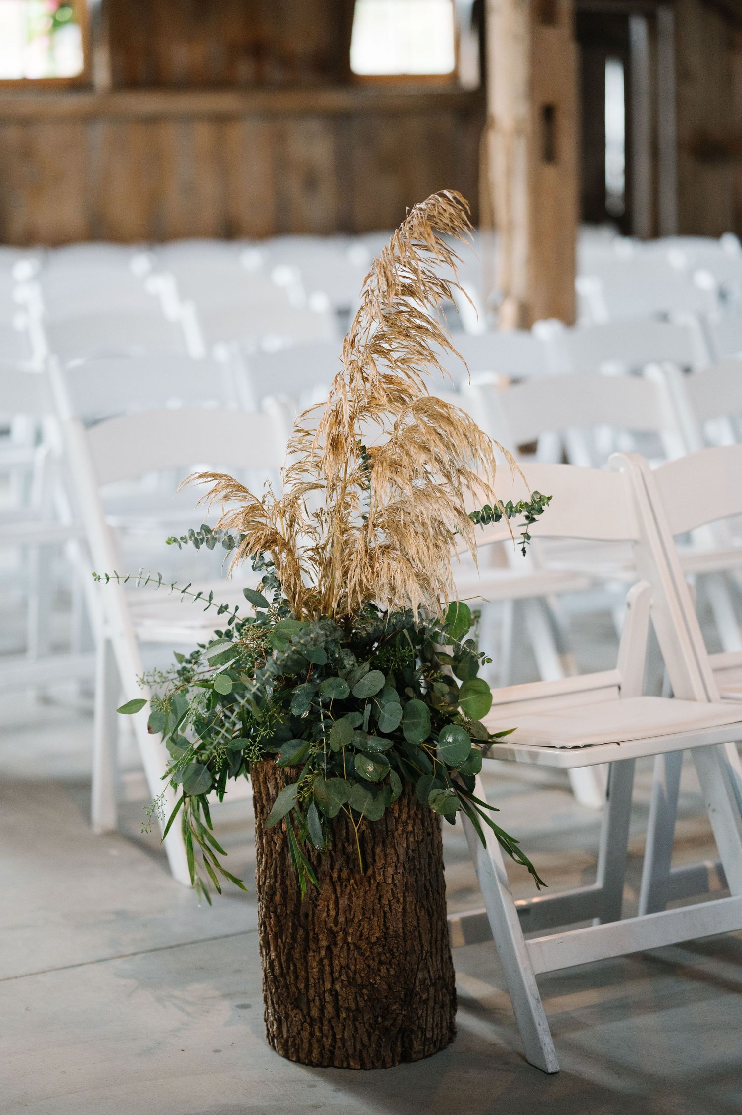 Rustic wedding ceremony decor featuring dried pampas grass and eucalyptus arranged on a log pedestal near white folding chairs.