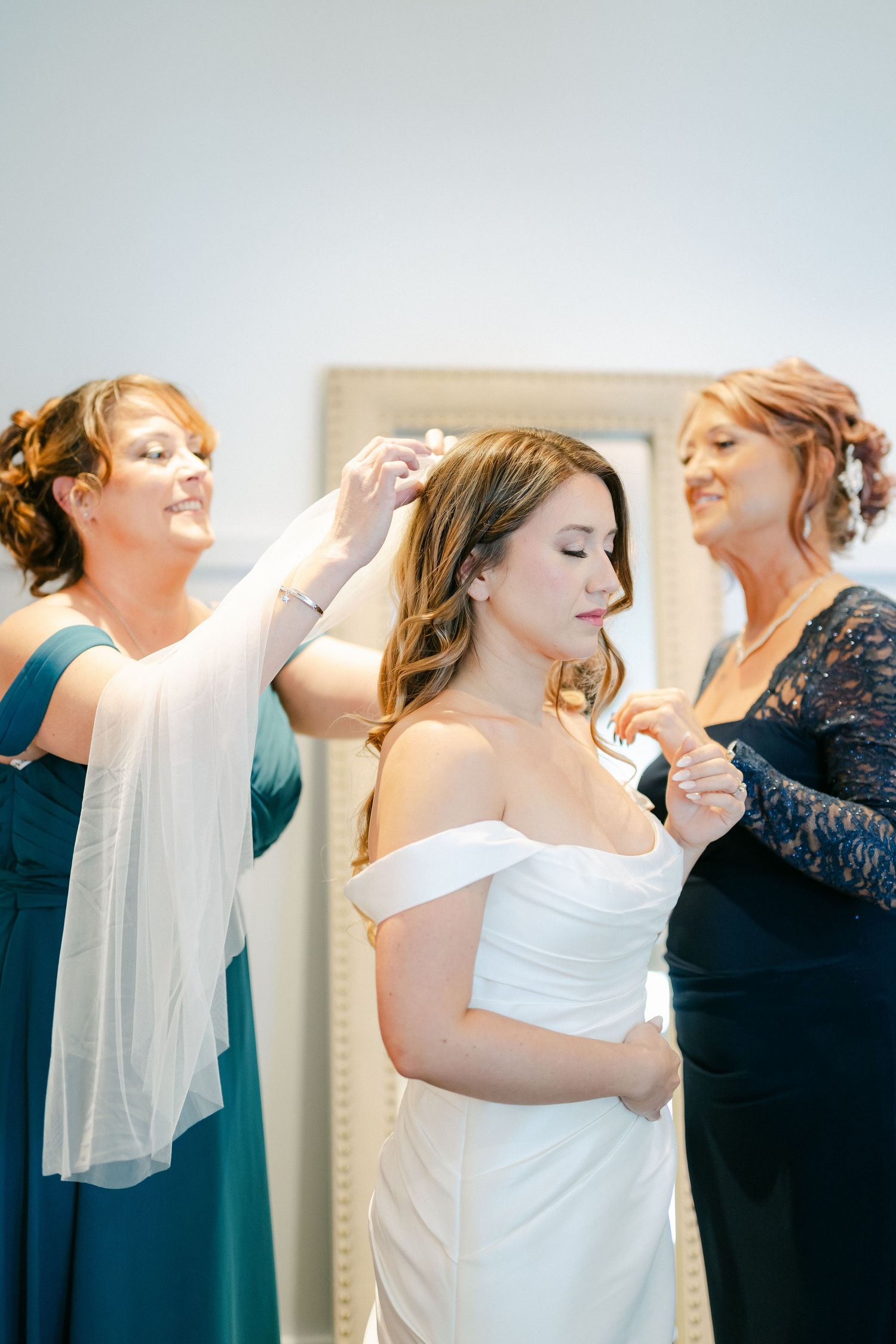 Bride gets help with final dress preparations from two women wearing blue and navy dresses.