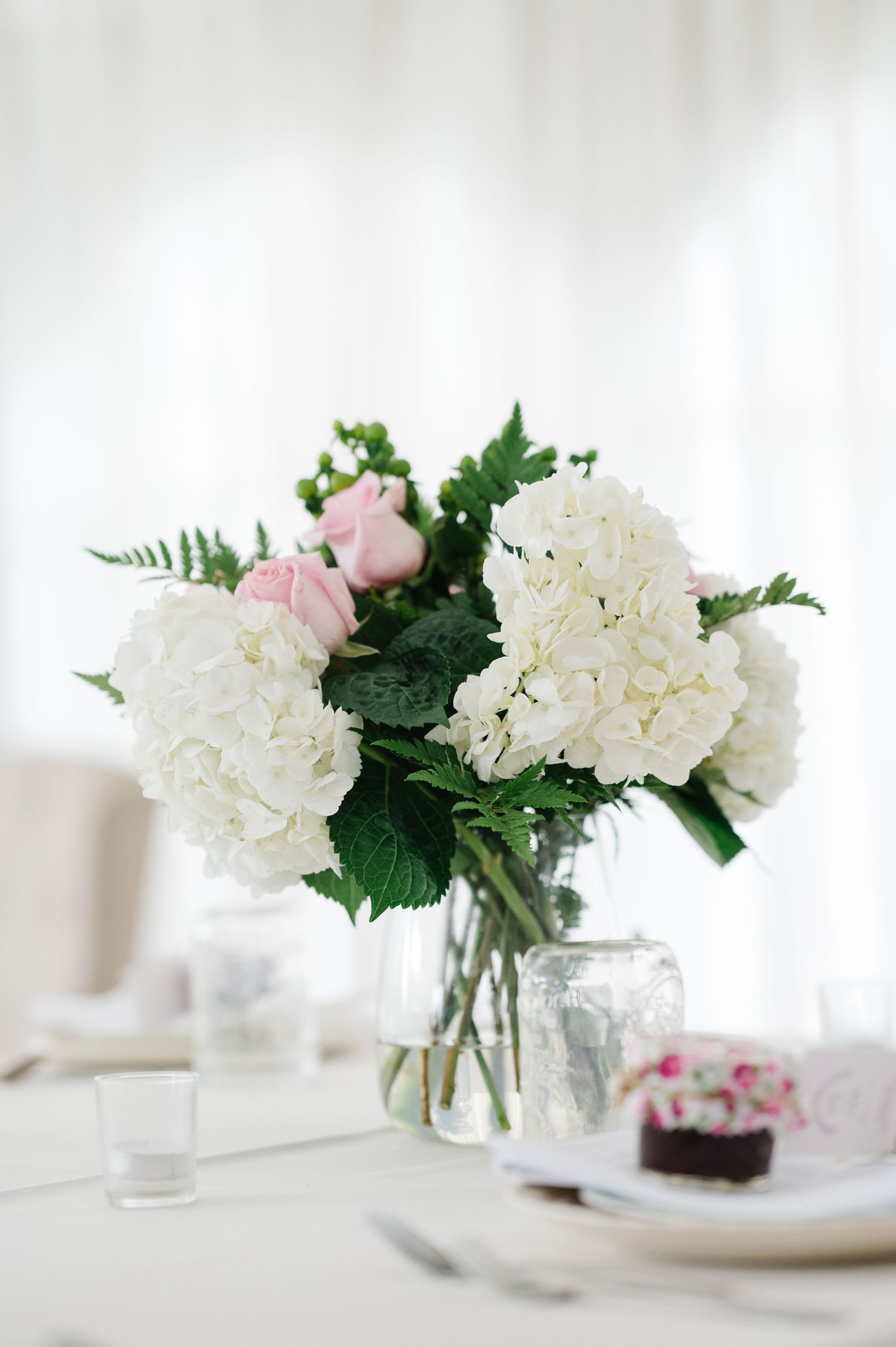 Elegant floral centerpiece with white hydrangeas and pink roses in a clear glass vase on white table setting.