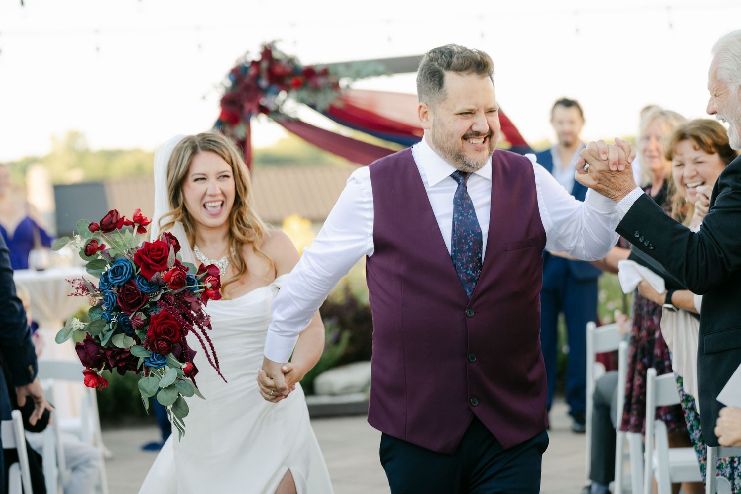 Joyful wedding couple walks hand in hand while exiting their ceremony with red floral bouquet and decorations.