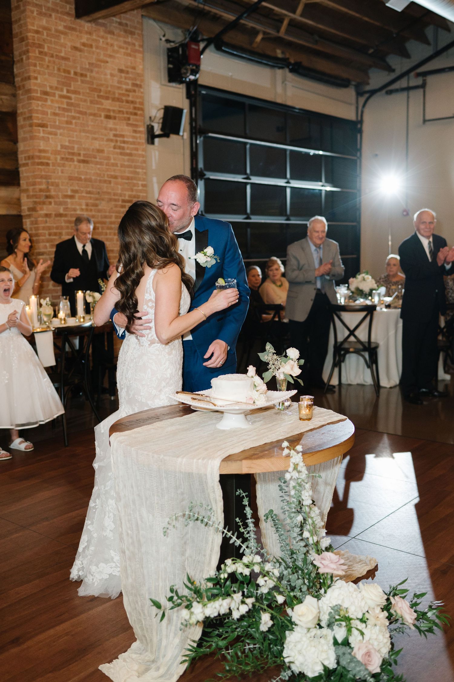A couple in wedding attire share a moment during their reception, with decorative floral arrangements and guests in background.