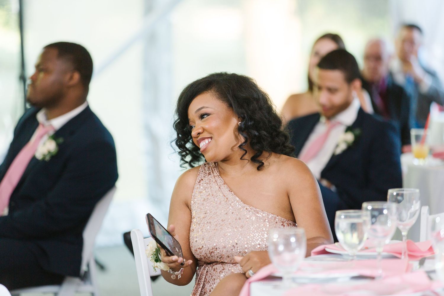Reception guest in a pink one-shoulder dress laughs while checking phone at wedding table decorated in pink accents.