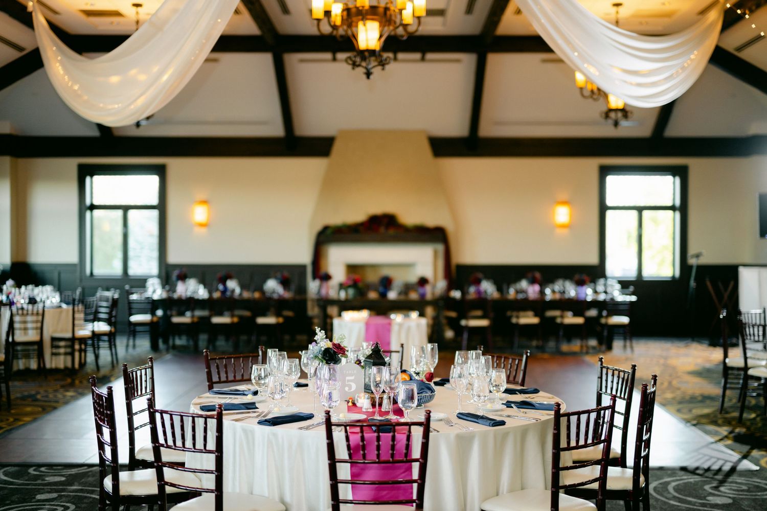 Elegant wedding reception venue with white linens, pink accent chairs and white draping decorations under vaulted ceiling.