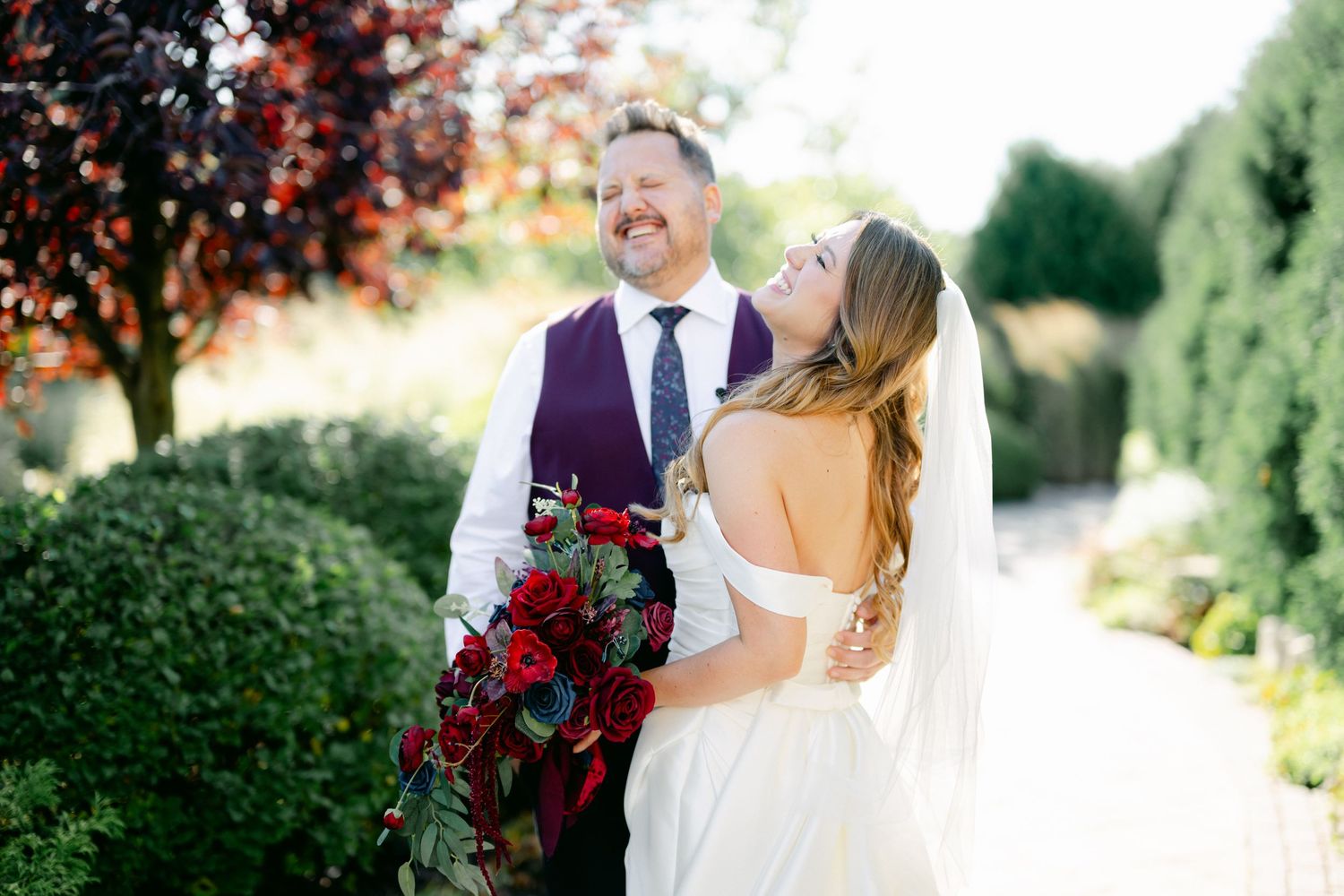 A couple in formal wedding attire poses in a garden setting with deep red floral bouquet and white bridal gown.