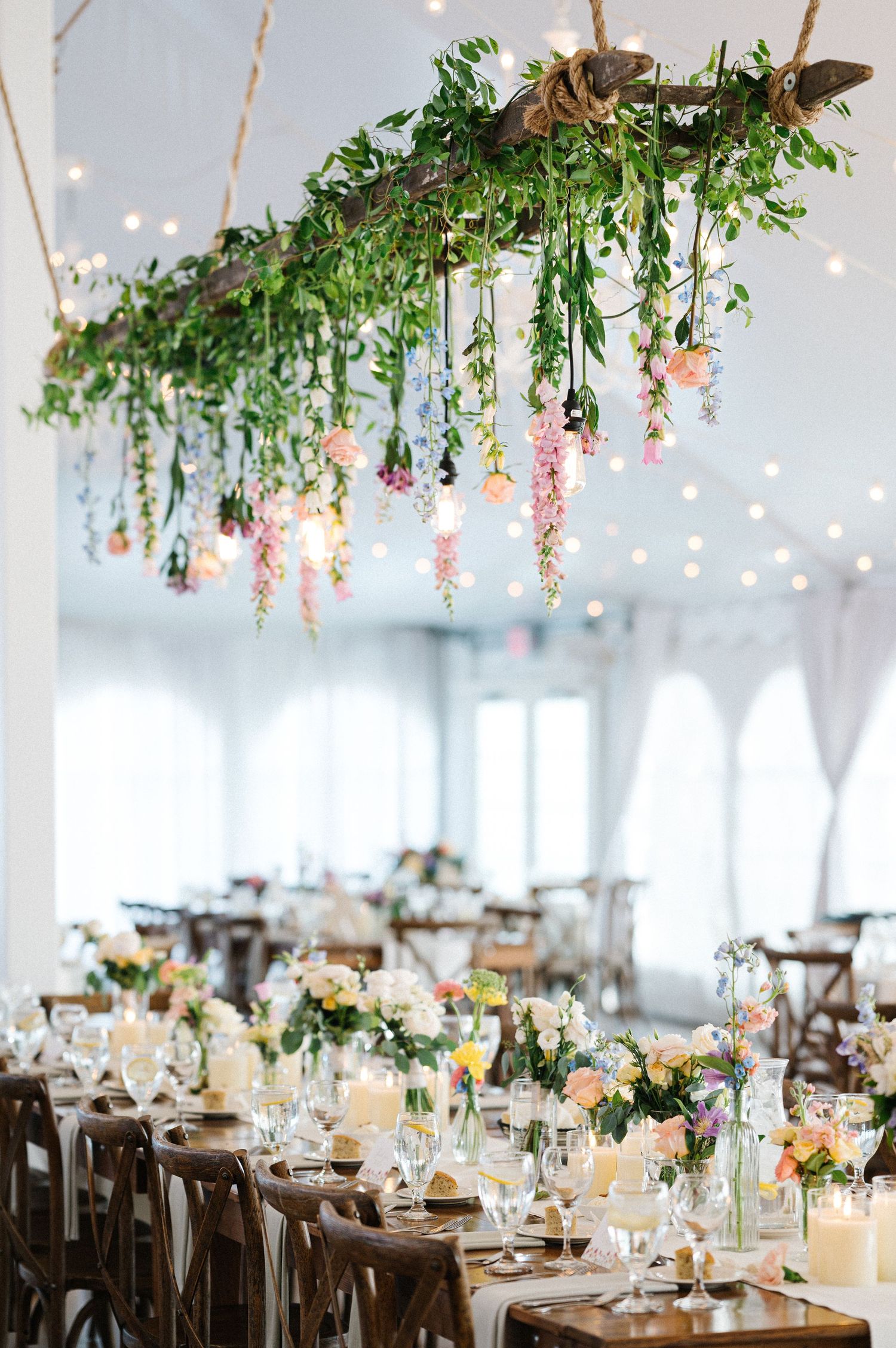 Elegant wedding reception setup with suspended greenery, pink flowers and twinkling lights over rustic wooden dining tables.