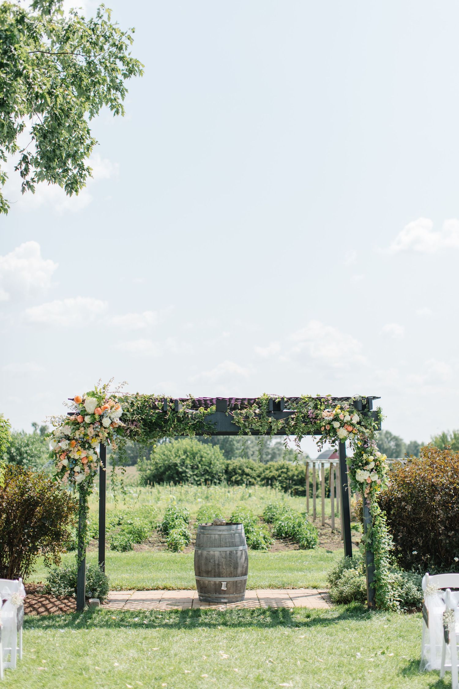 Rustic wedding arch decorated with flowers and greenery stands in a garden setting with a wooden barrel centerpiece.