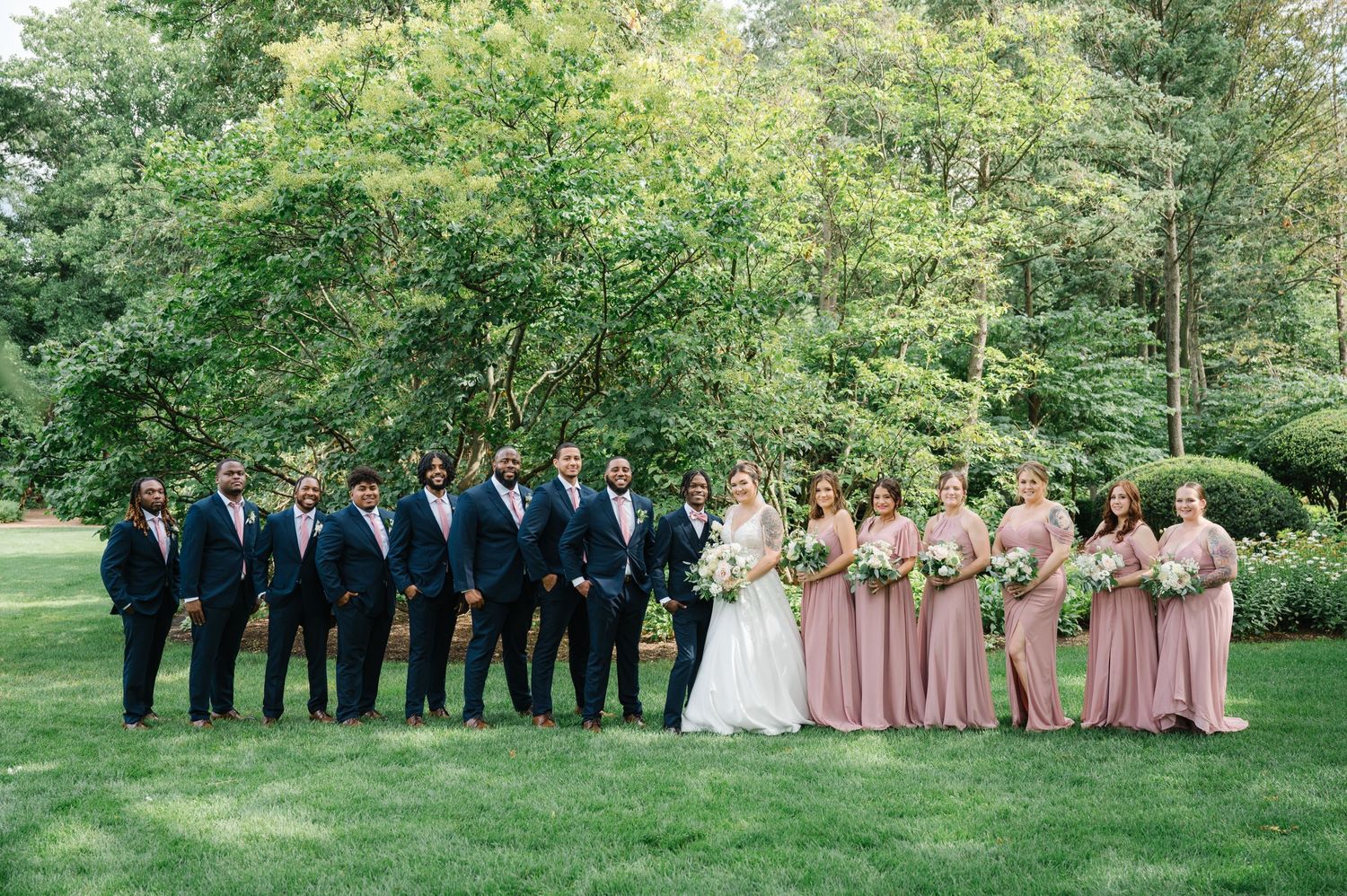 A wedding party in navy suits and blush pink dresses poses together on a lush green lawn surrounded by trees.