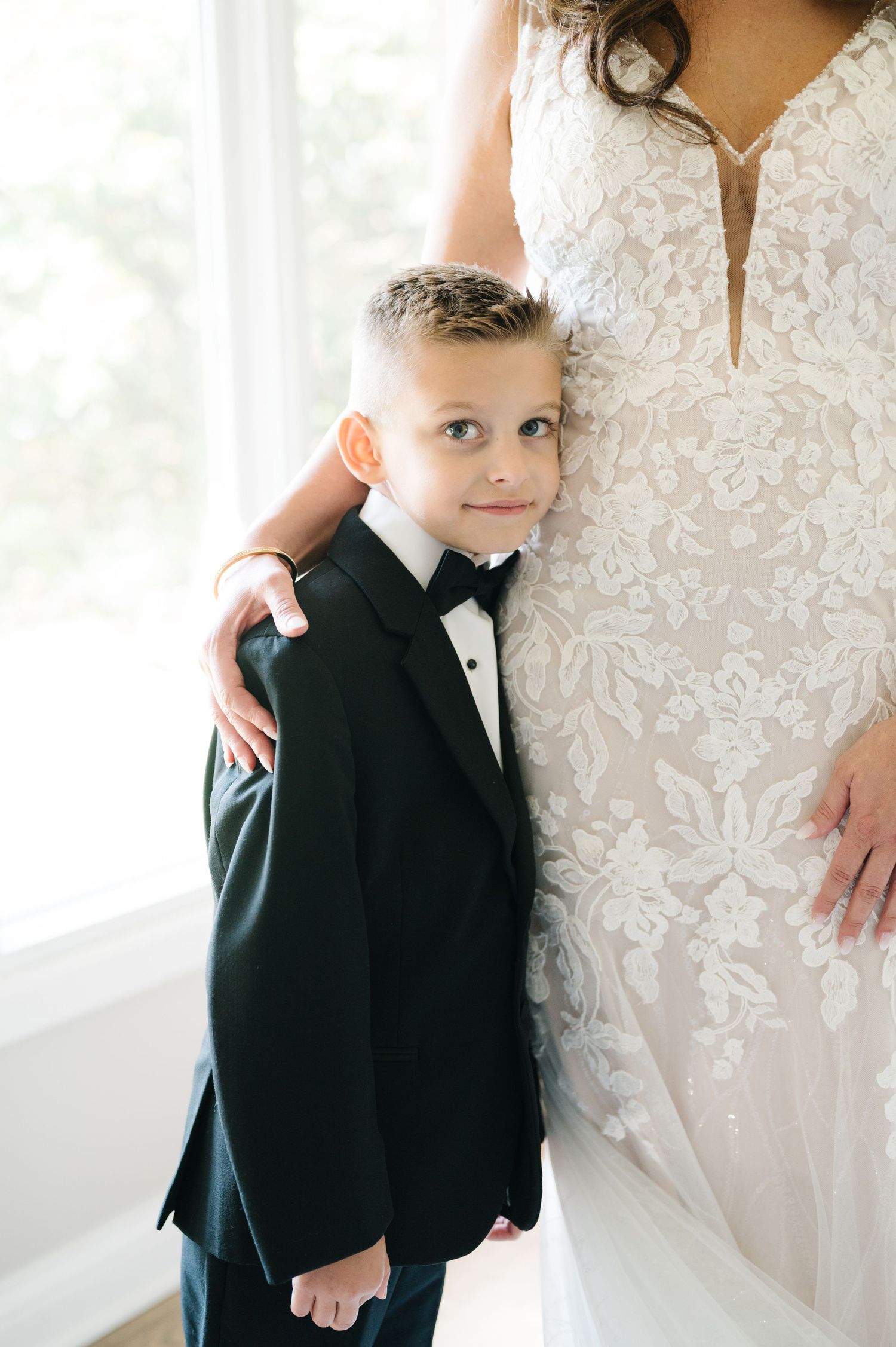 Small child wearing a black tuxedo with bowtie stands next to a white lace wedding dress in a bright indoor setting.