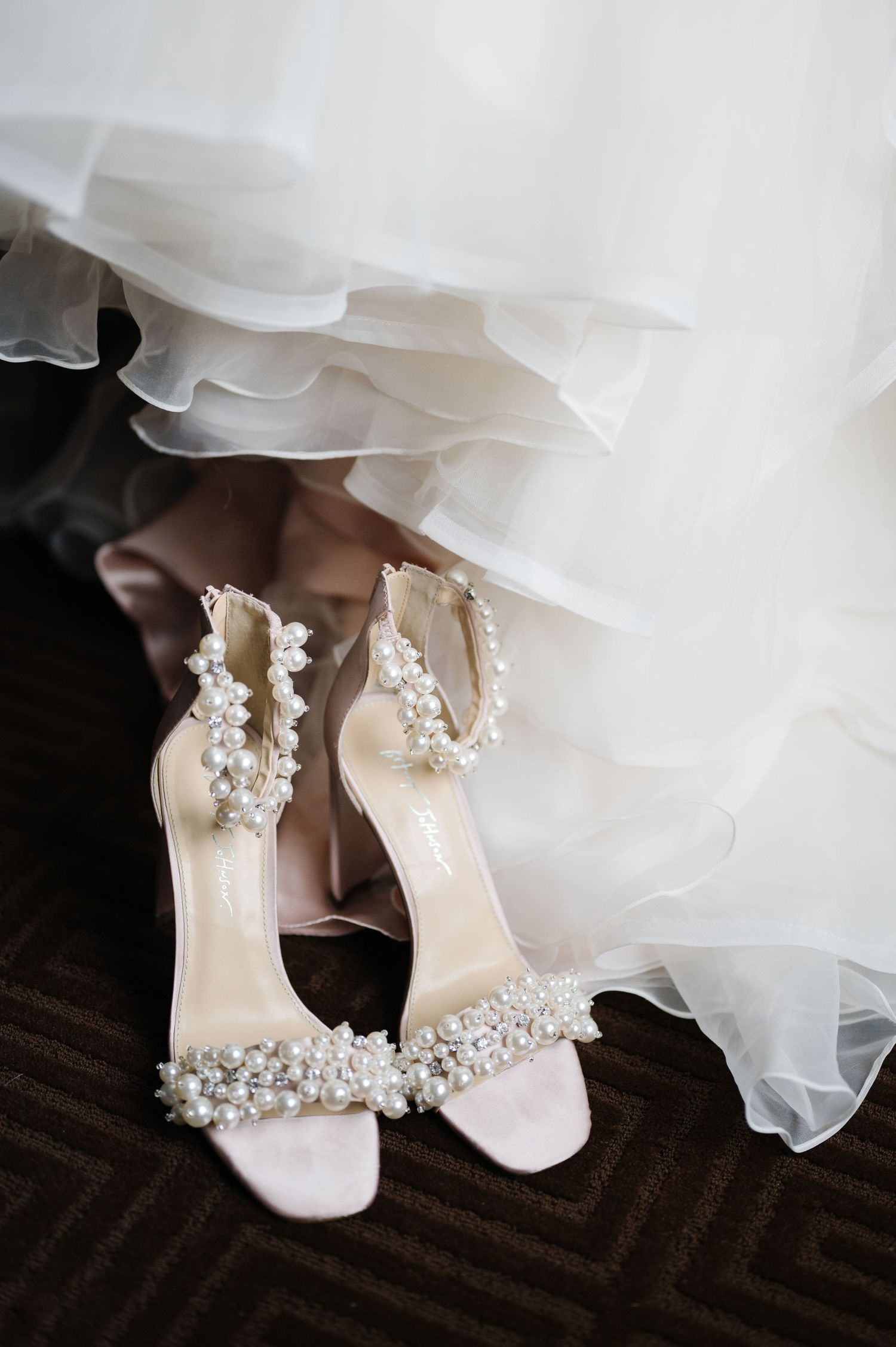 Elegant bridal shoes adorned with pearl embellishments peek out beneath layers of flowing white tulle veil.