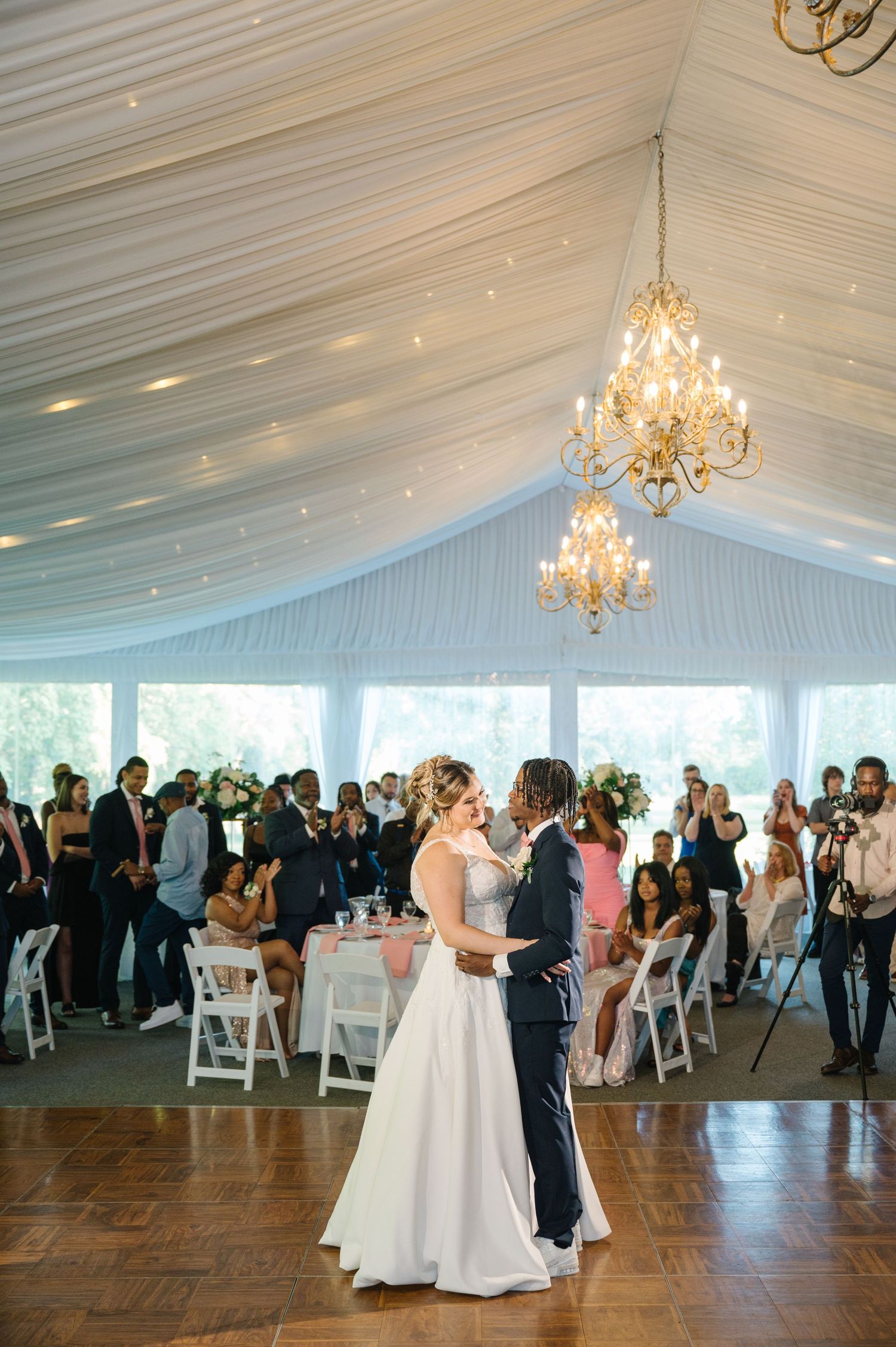 A couple shares their first dance under elegant crystal chandeliers in a white tent-draped reception venue.