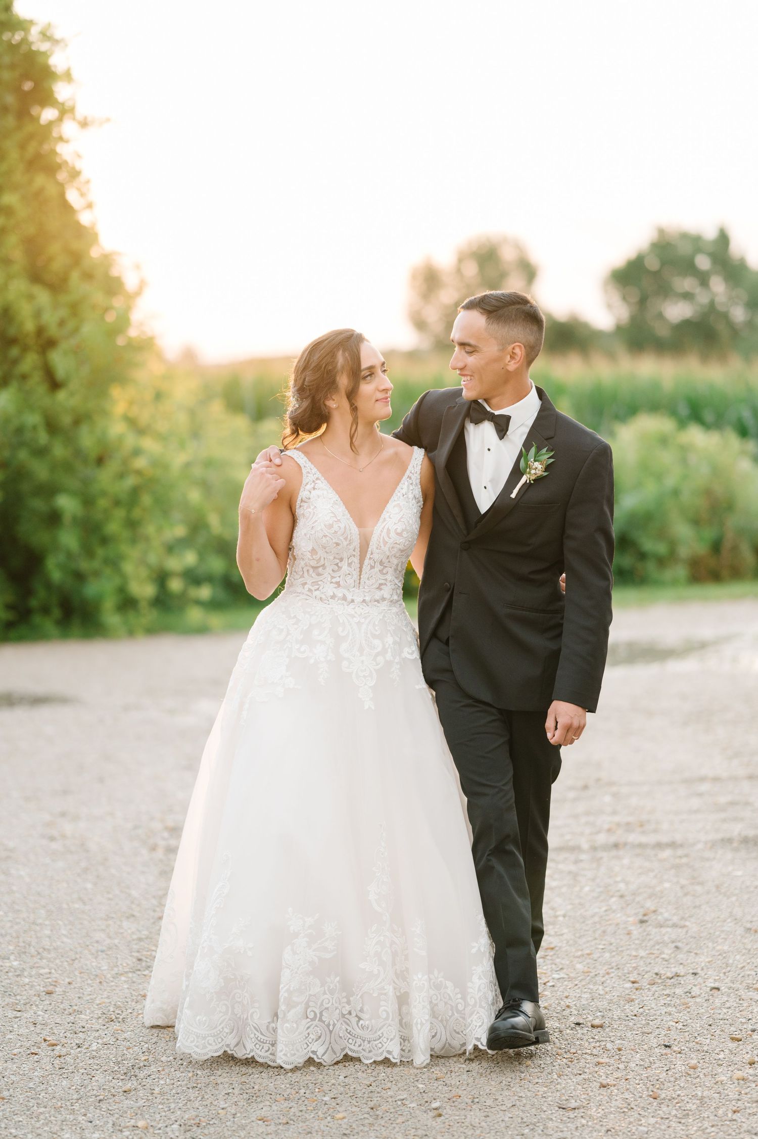 A bride in a lace ball gown and groom in classic tuxedo walk together on a path during golden hour.