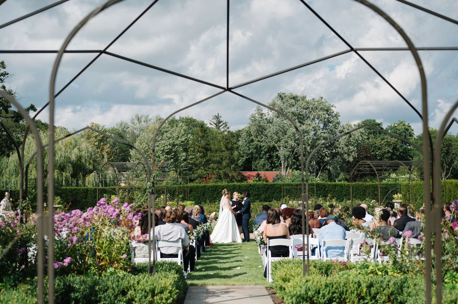 Outdoor garden wedding ceremony under a metal arch structure with guests seated among pink flowers and lush greenery.