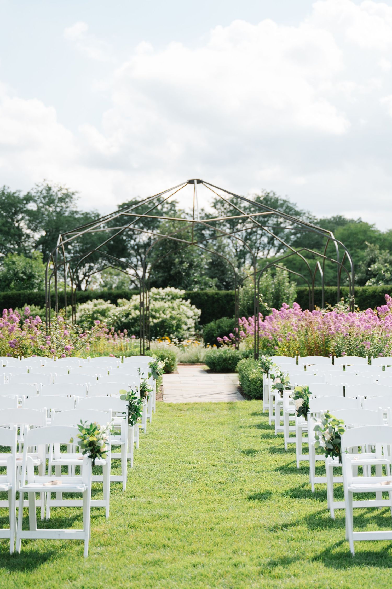 Outdoor wedding ceremony setup with white chairs on grass and floral arch decorated with pink and white flowers.
