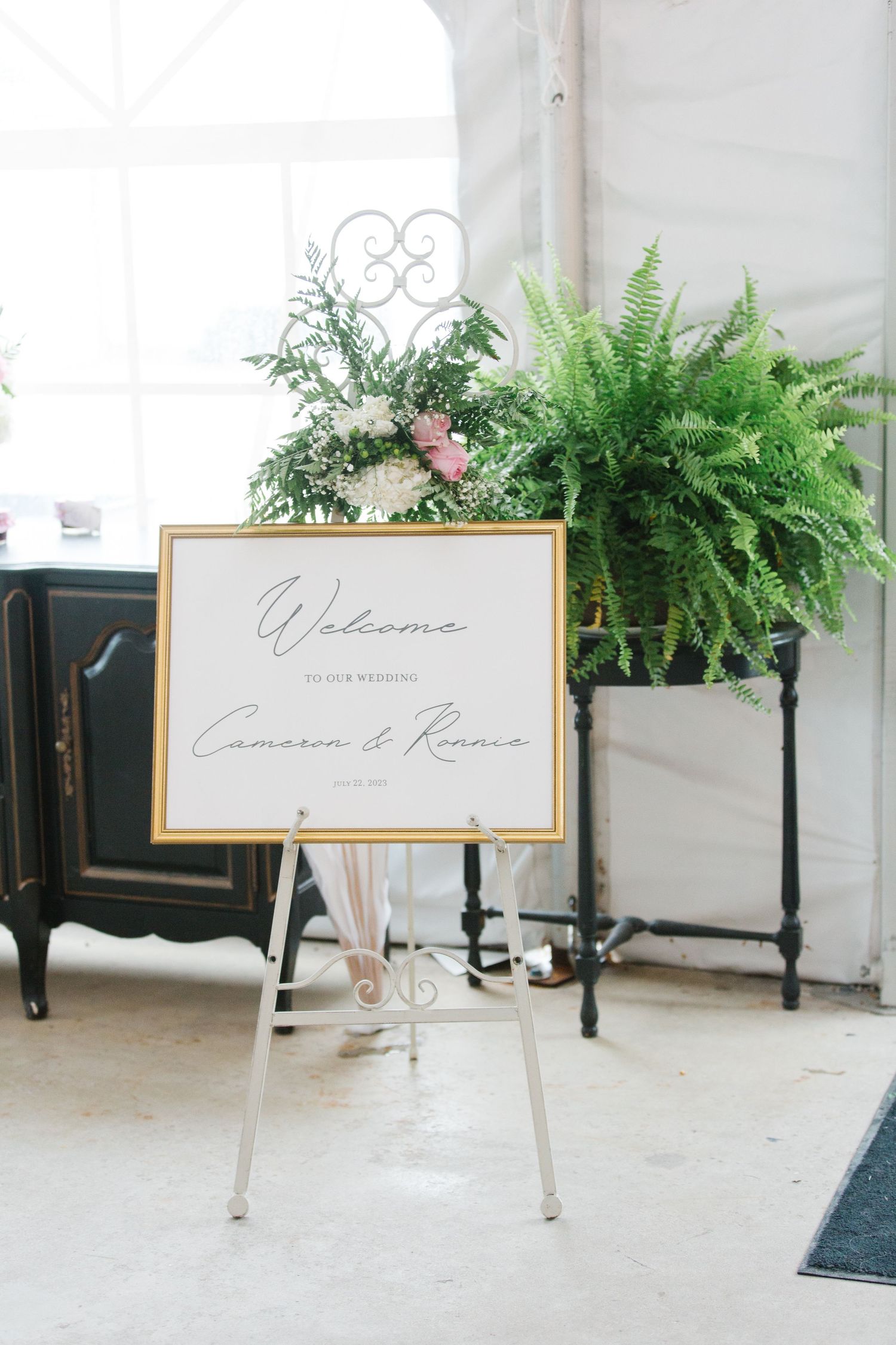 A welcome sign on an easel stands next to potted ferns in a bright white room with minimal decor.