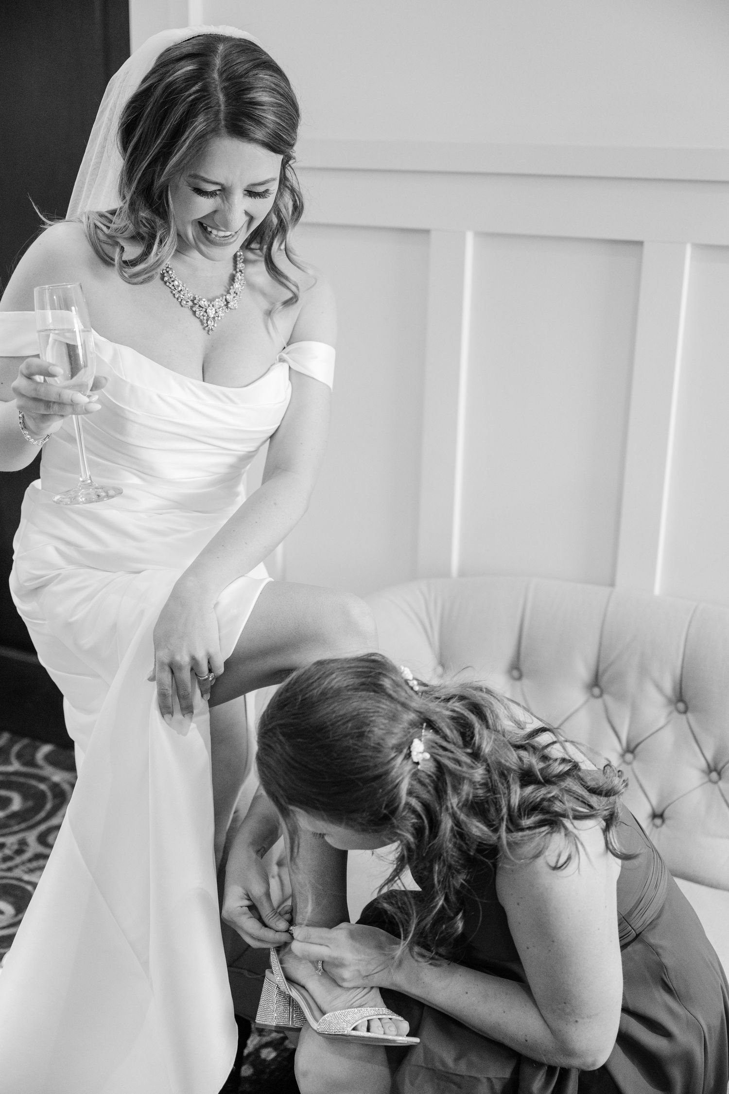 At a wedding preparation, someone in an off-shoulder white gown holds a drink while another person works on a shoe.