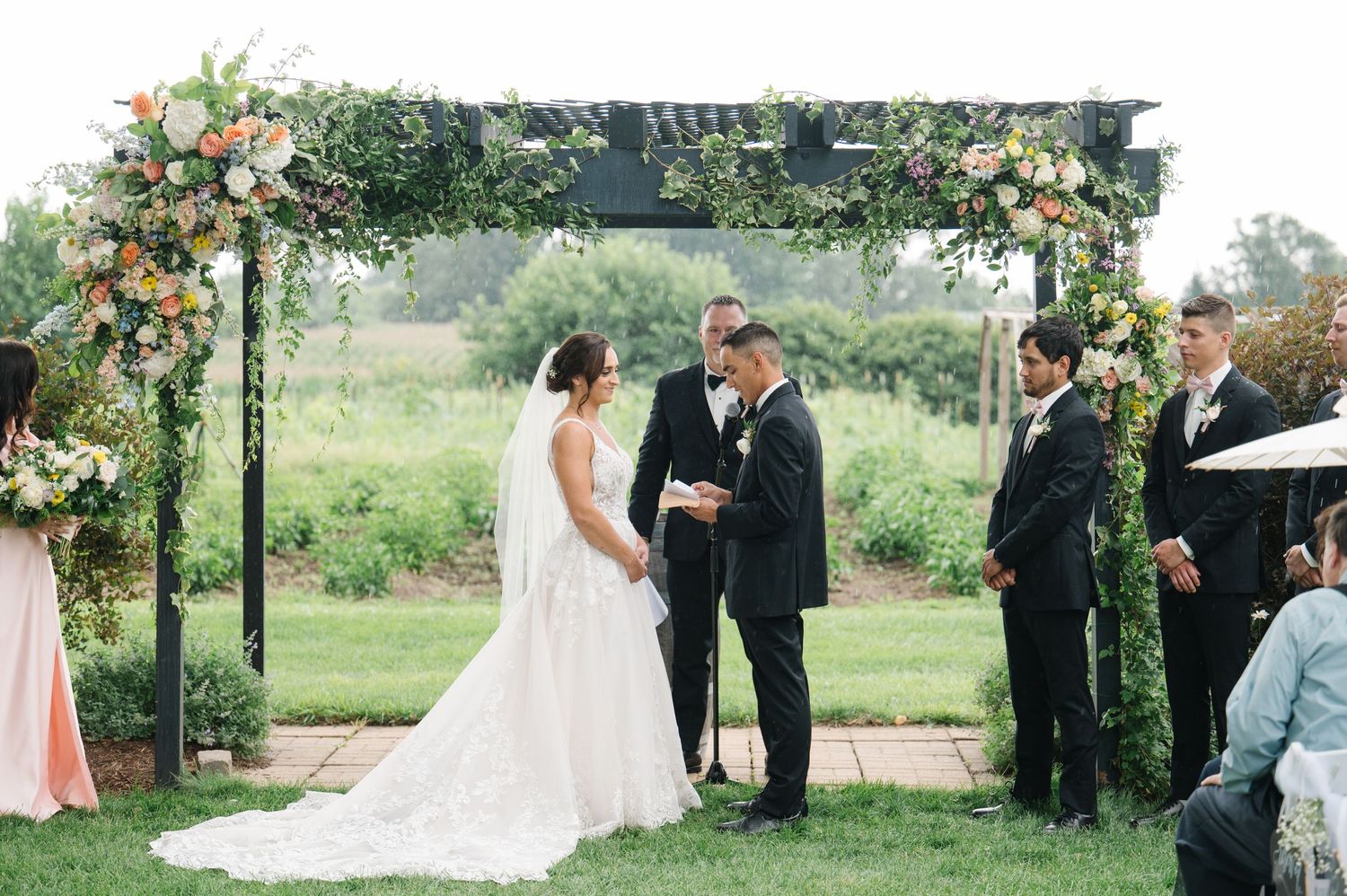 An outdoor wedding ceremony takes place under a floral arch with pink and peach blooms in a lush garden setting.