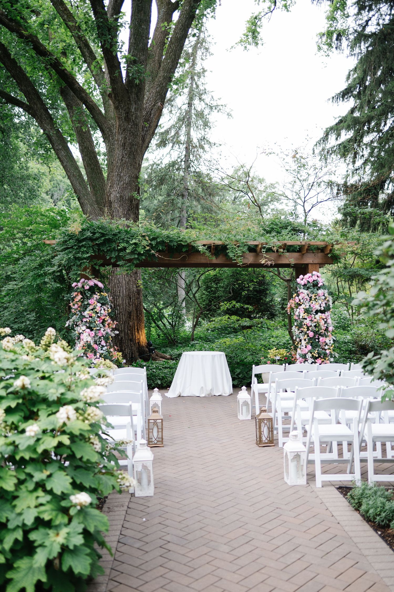 Outdoor garden wedding ceremony setup with white chairs, floral arch, and lanterns along a brick path under tall trees.