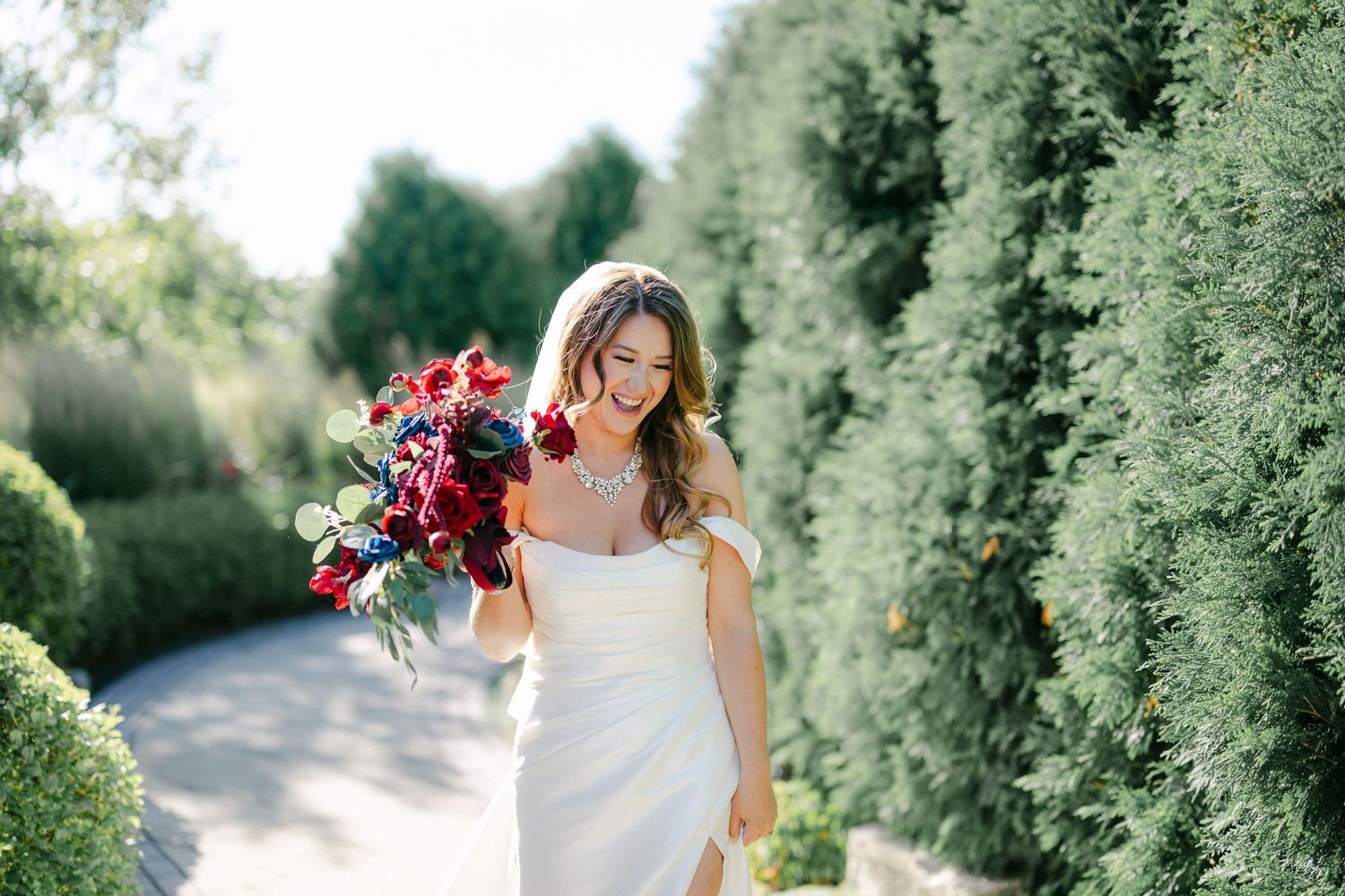 A bride in a white wedding dress holding a vibrant red and blue bouquet walks along a garden path.