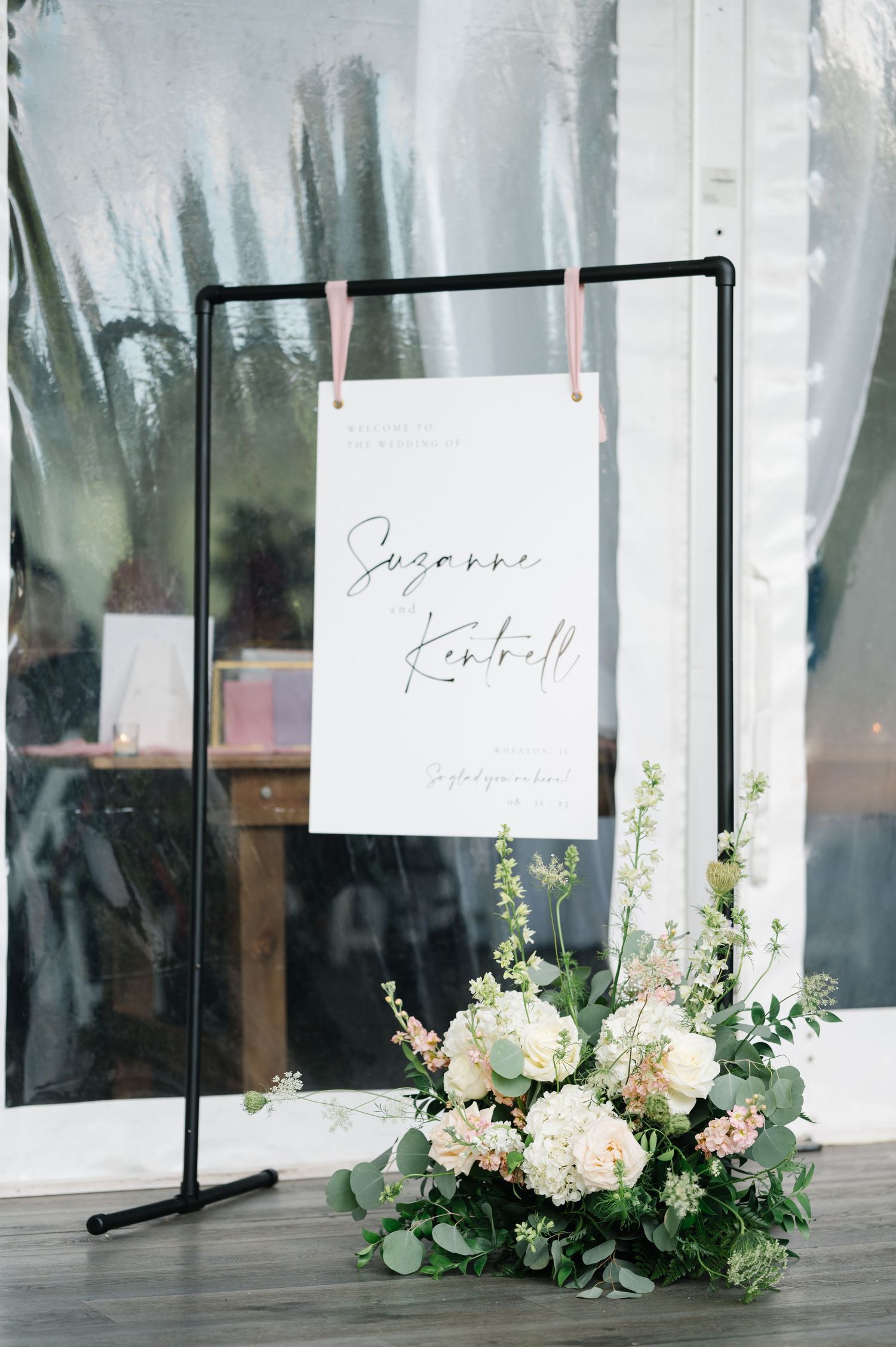 Elegant wedding signage display with white handwritten text on black stand, adorned with soft pink and white floral arrangement.