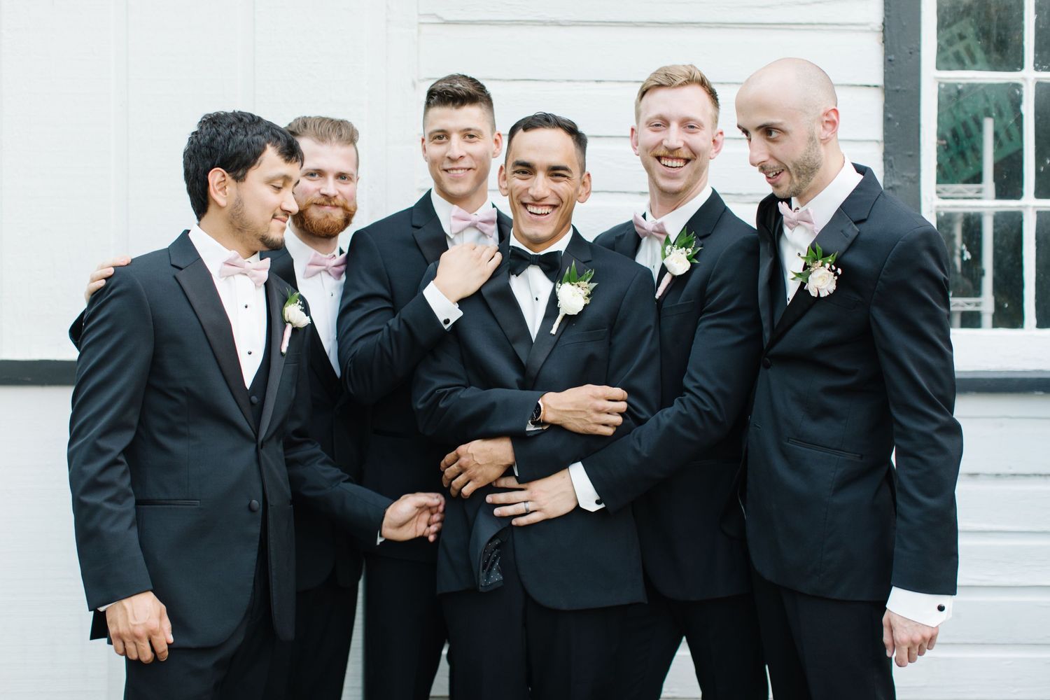 A group in formal black tuxedos and suits smile while standing together outside a white building.