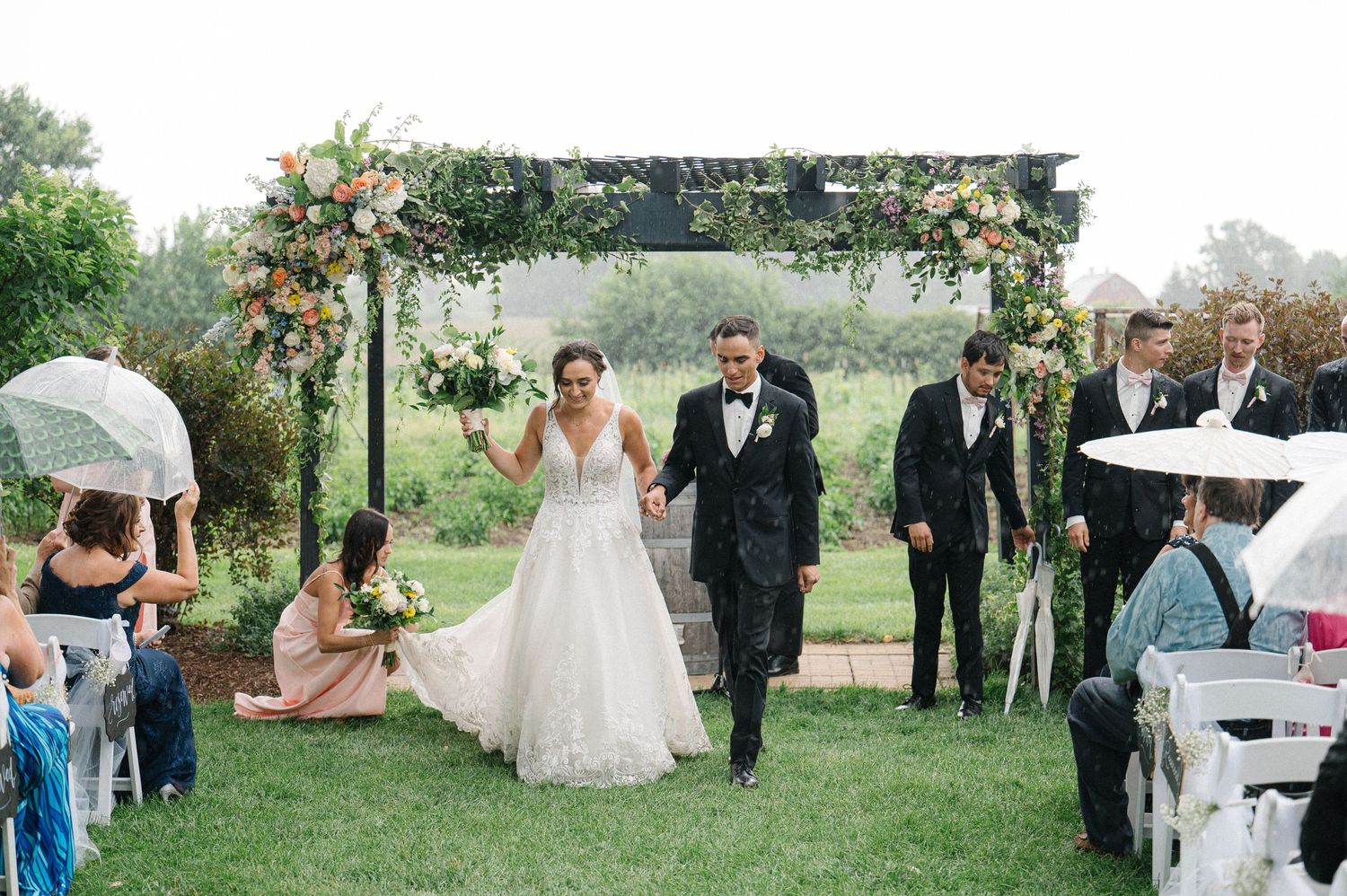 A wedding ceremony takes place outdoors under a floral arch with guests seated on the lawn holding umbrellas on a misty day.