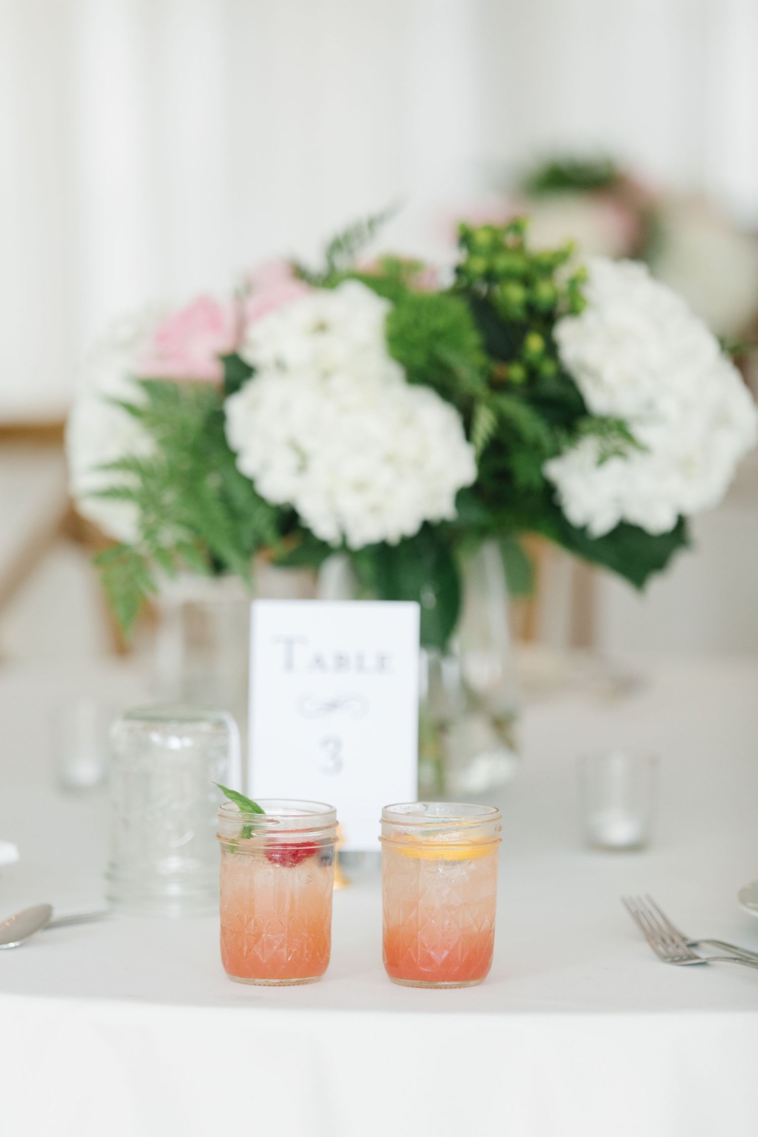 Elegant table setting with mason jar cocktails and white hydrangea floral centerpiece on white linen tablecloth.