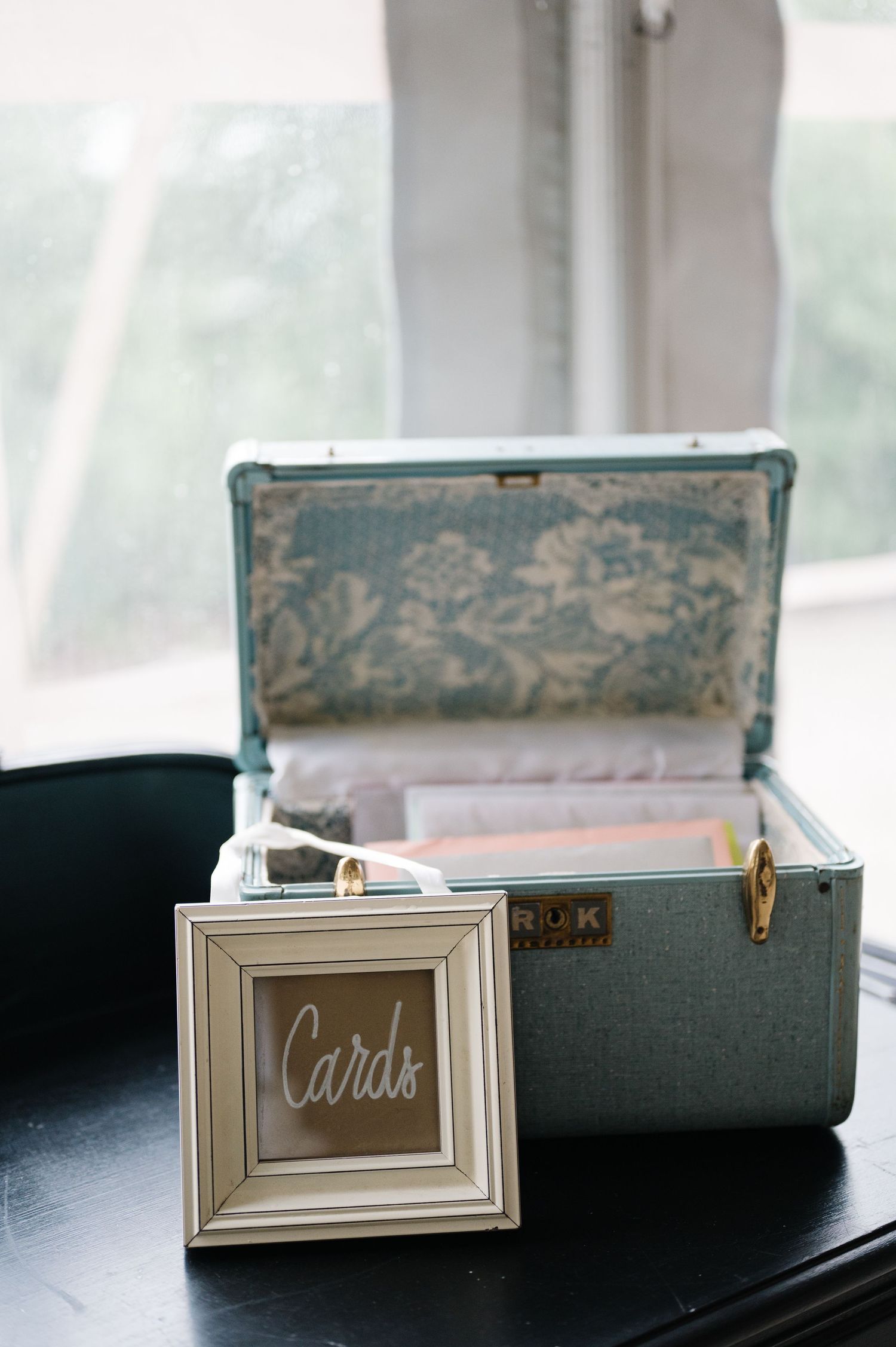 Vintage suitcase with decorative floral lining next to small framed 'Cards' sign on dark table.