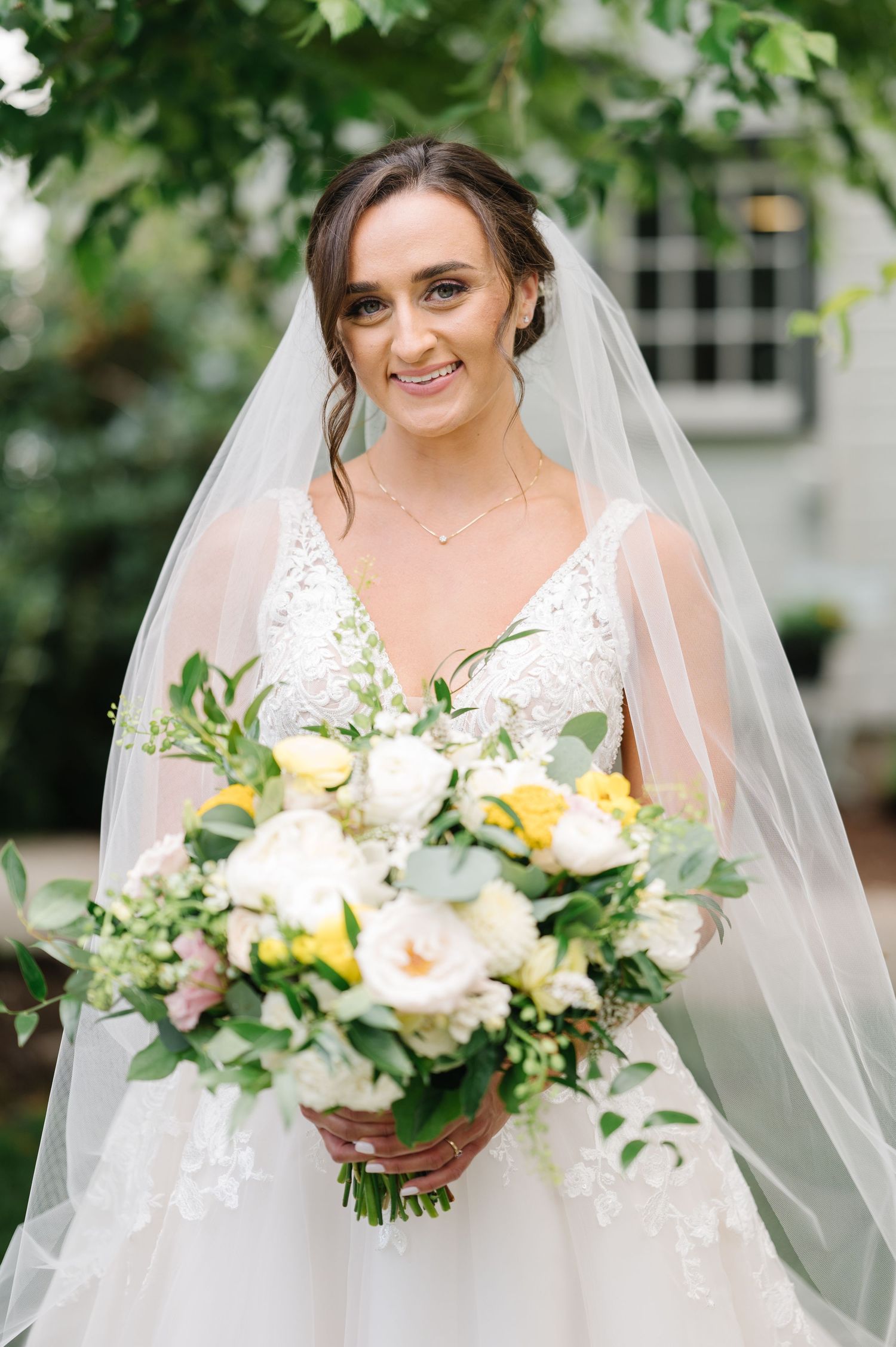 A bride in a white lace wedding dress holds a beautiful spring bouquet with yellow and white flowers and greenery.