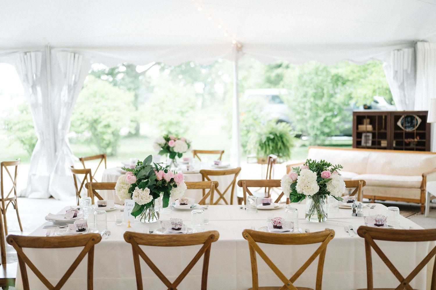 Elegant wedding reception table setting with wooden cross-back chairs and white floral centerpieces under a tented venue.