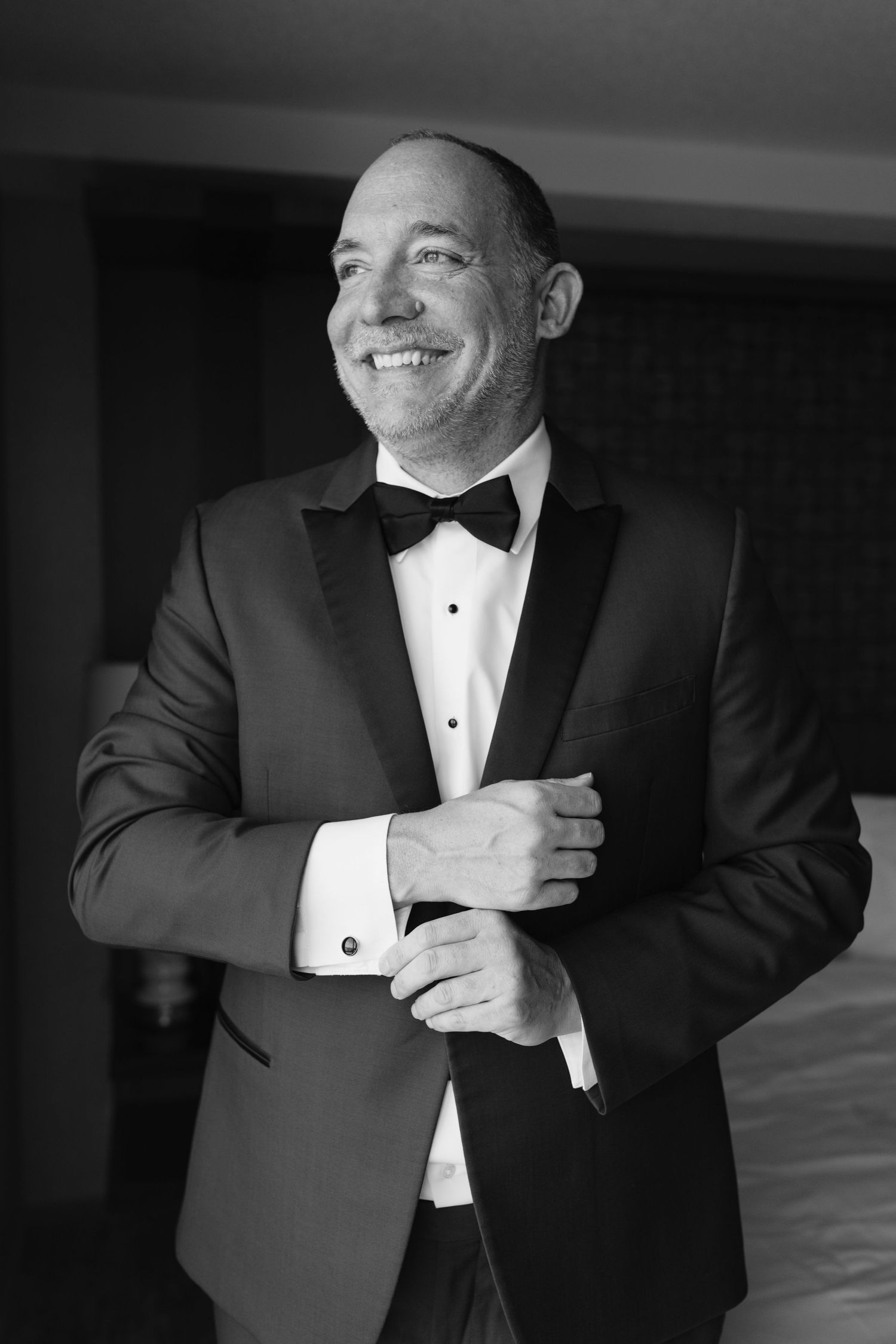 Black and white portrait of person in tuxedo with bow tie adjusting their cuff and smiling.