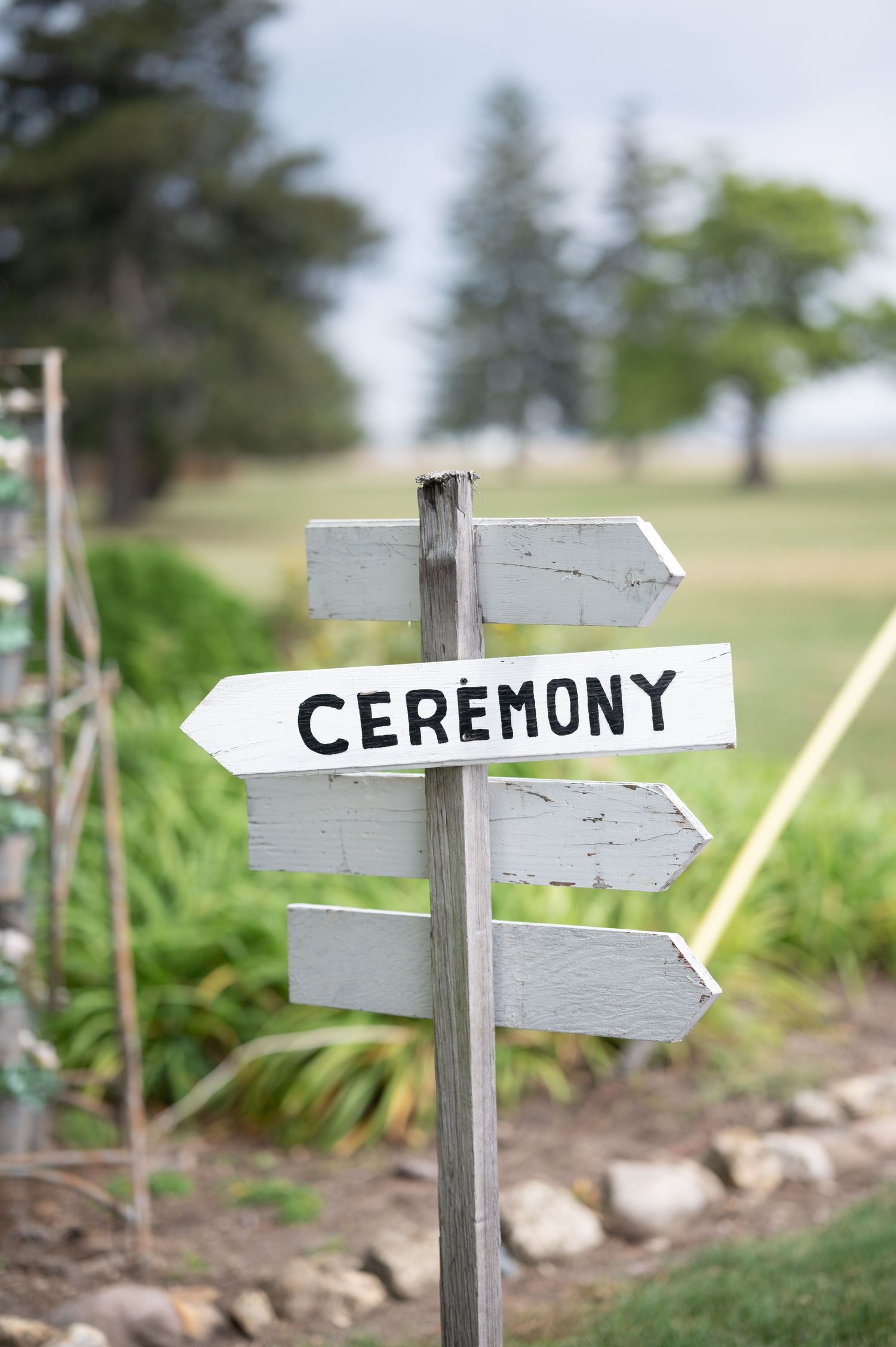A rustic wooden directional sign with the word 'Ceremony' pointing to the right against a blurry natural background.