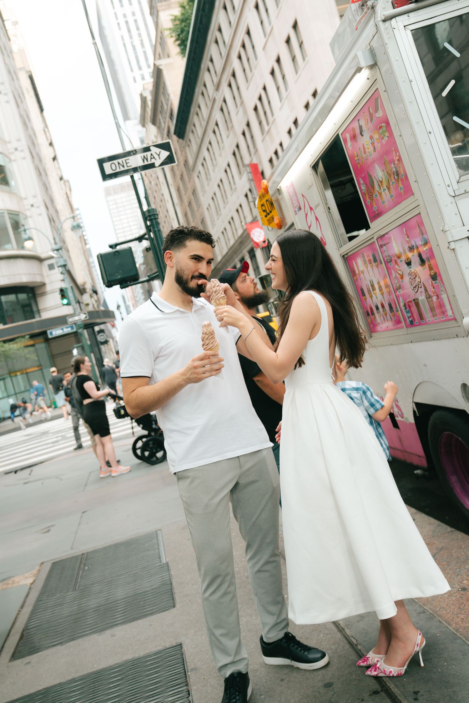 A couple enjoys ice cream cones while standing on a city street corner on a sunny day.