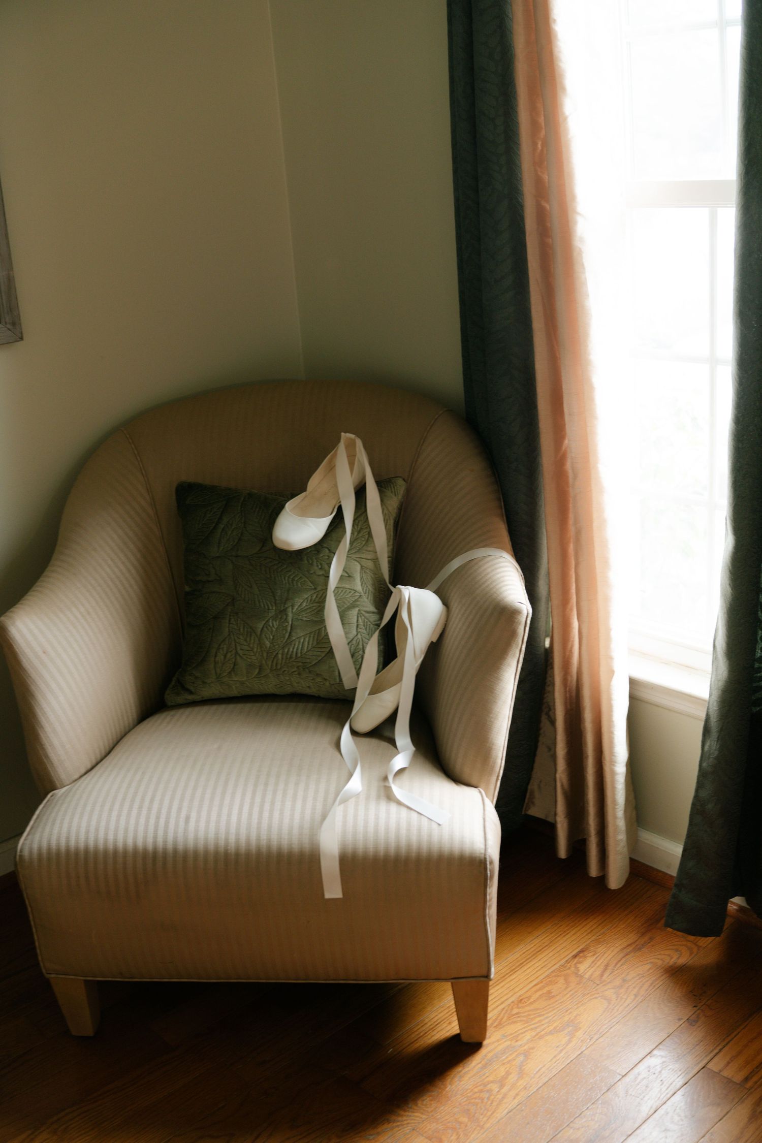 Elegant white high heel shoes placed on a beige upholstered armchair with green cushion by a curtained window.