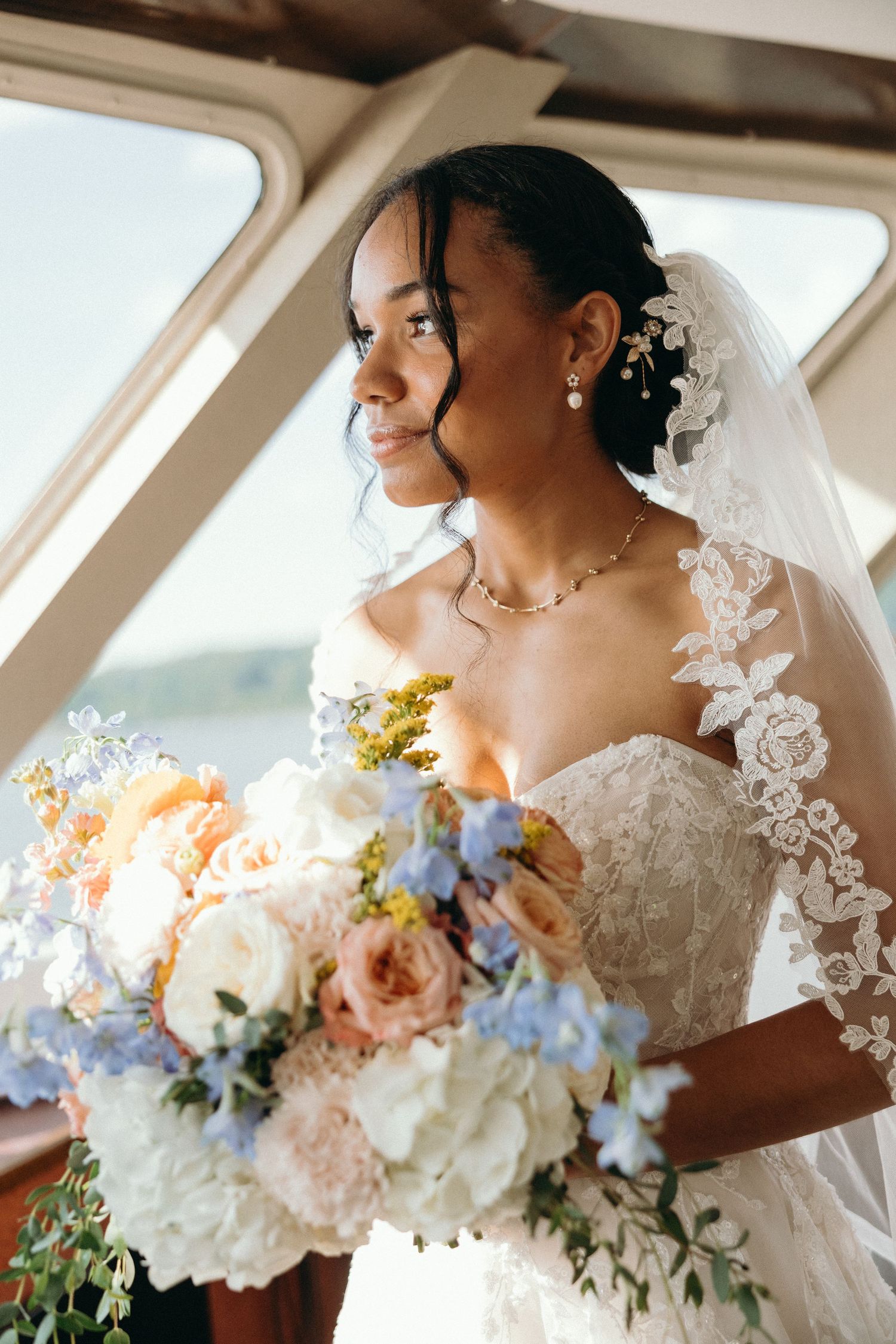 Bride in strapless lace wedding gown holds elegant pastel bouquet with peach roses, white peonies and blue delphinium flowers.