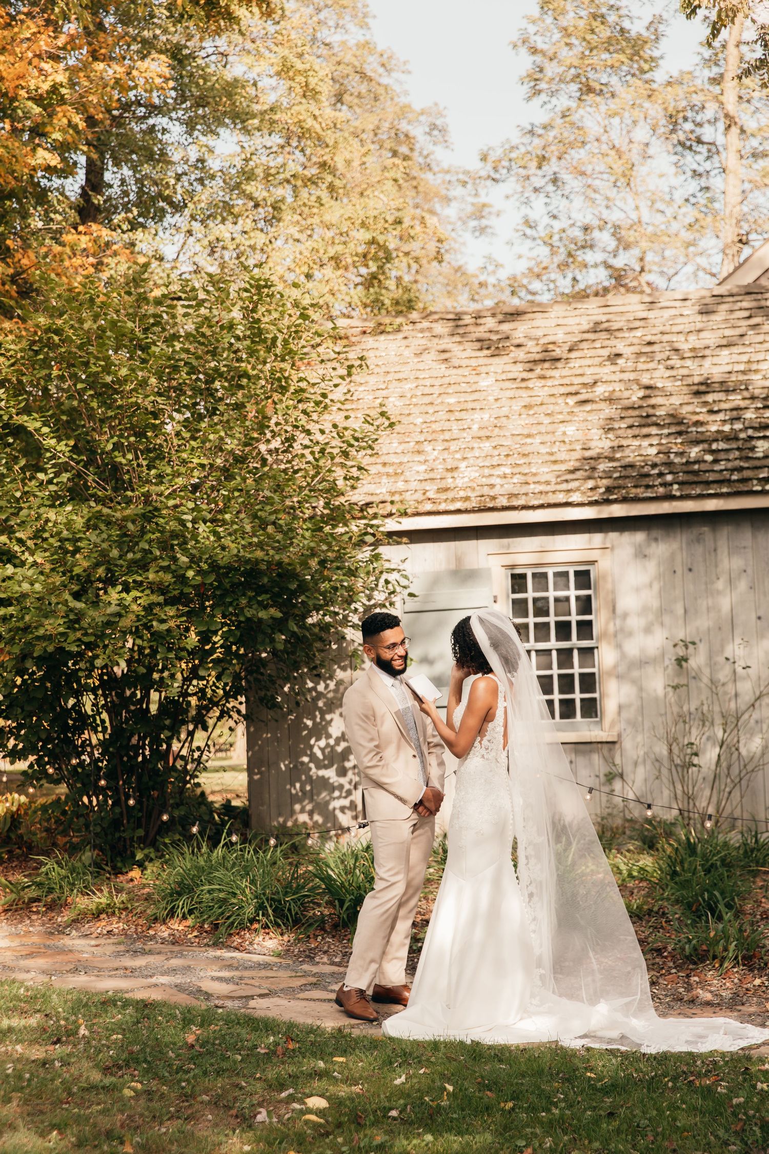 A couple stands together on an autumn day outside a rustic wooden building with fall foliage in the background.