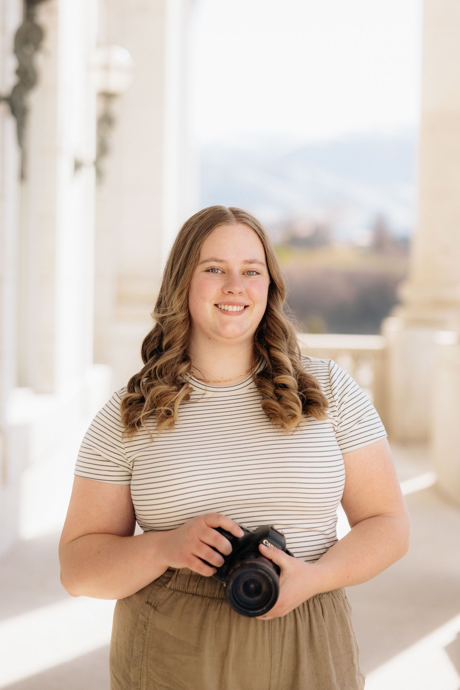 Young photographer in striped t-shirt and brown skirt holds a camera while standing in bright outdoor corridor.
