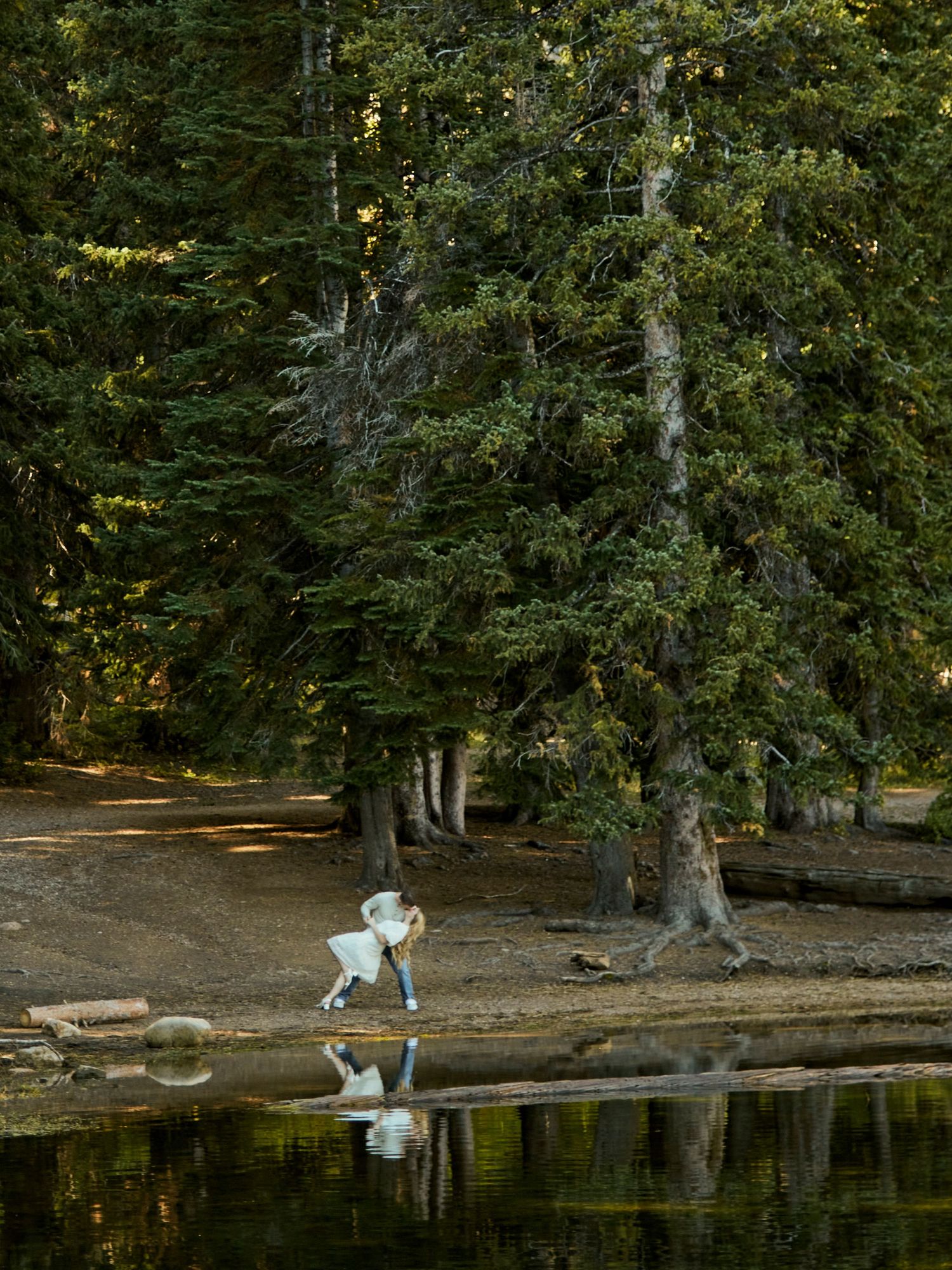 Person in blue outfit reflected in calm lake water surrounded by tall evergreen trees.