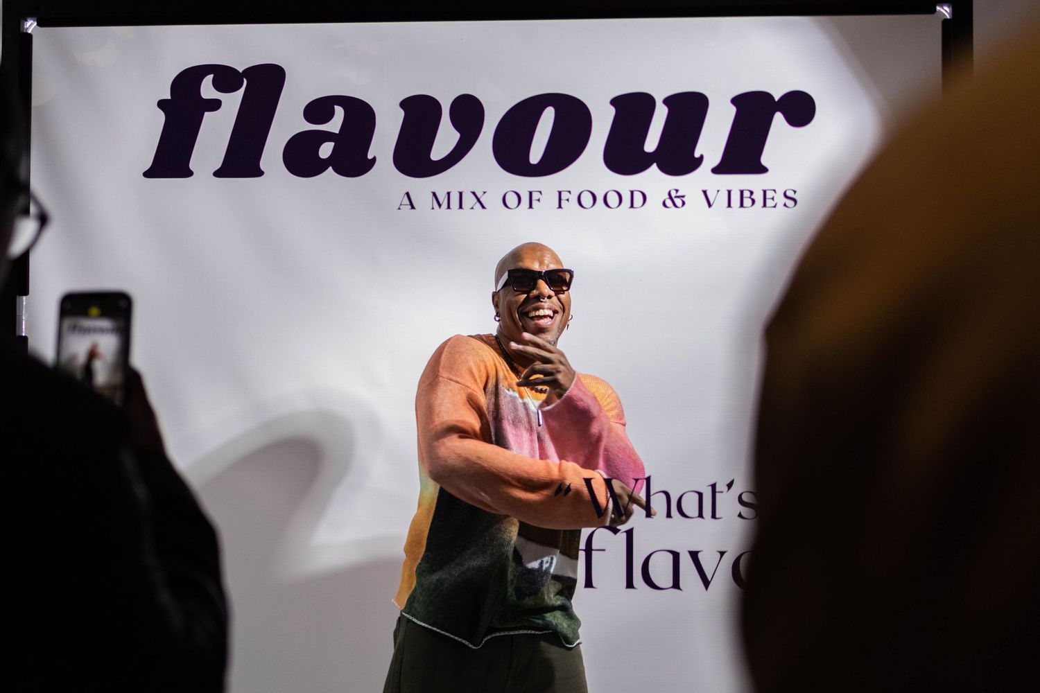 Person performs on stage with 'flavour' text display behind them in dark lighting.