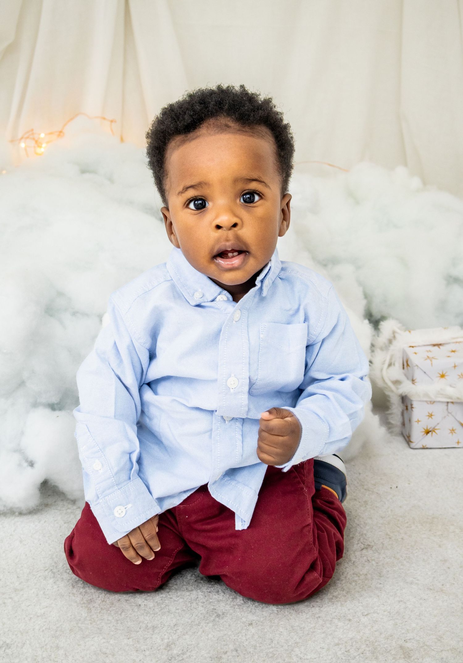 Young toddler in light blue shirt and burgundy pants sitting on fluffy white backdrop with holiday decor.