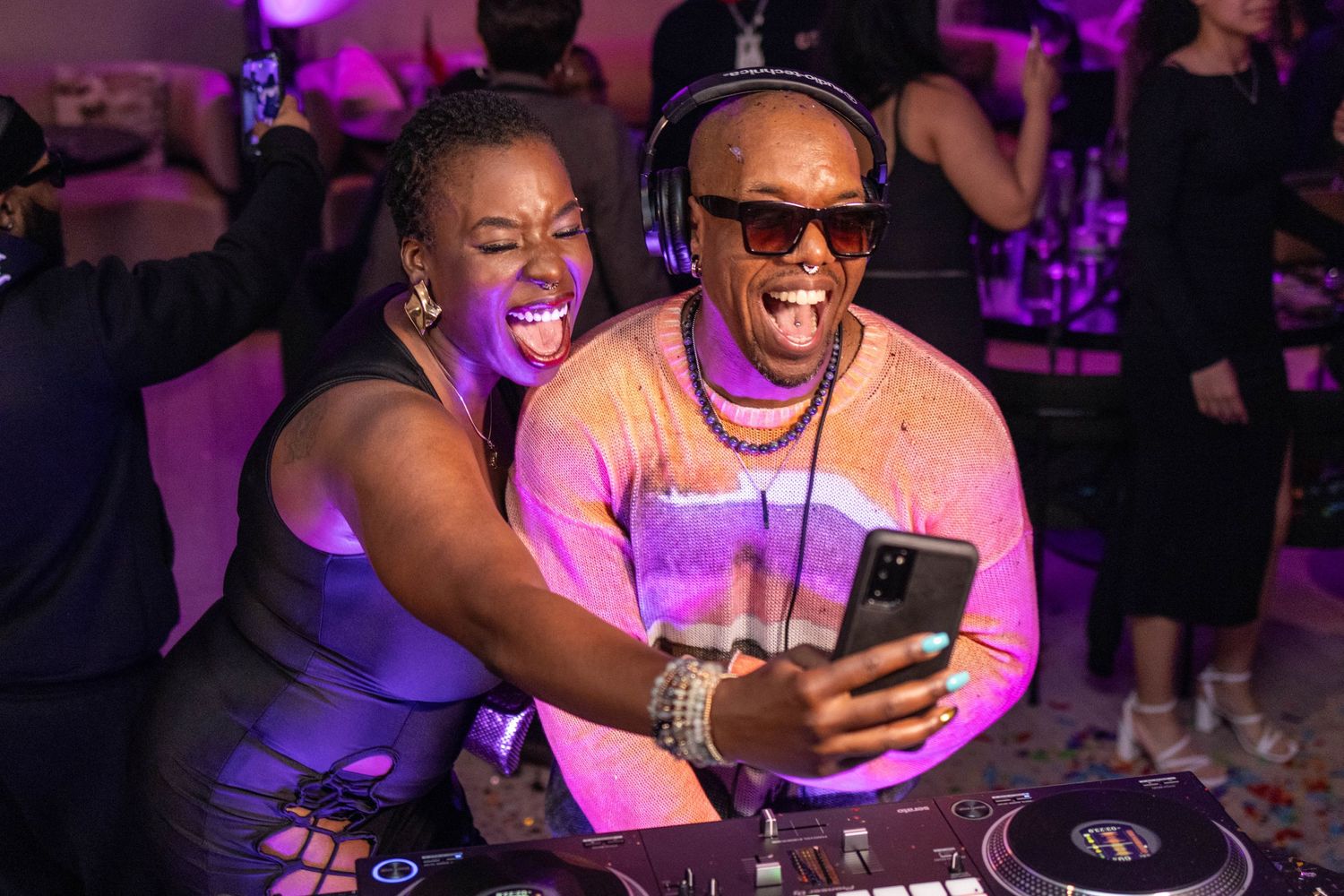 DJs in bright pink lighting share a moment while looking at a phone during a nightclub event.