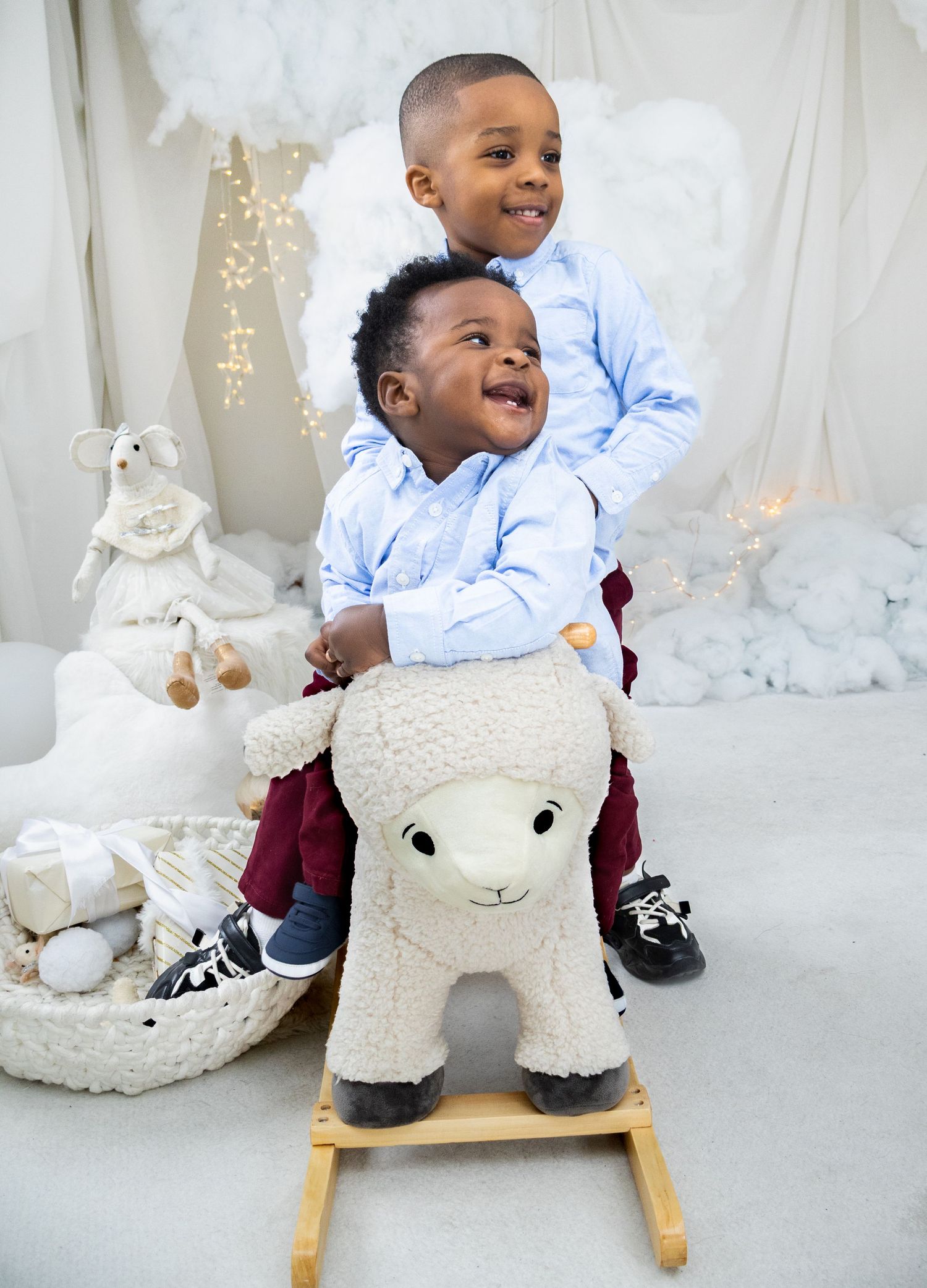 Two children in light blue shirts share joyful smiles while riding a plush white rocking sheep toy.