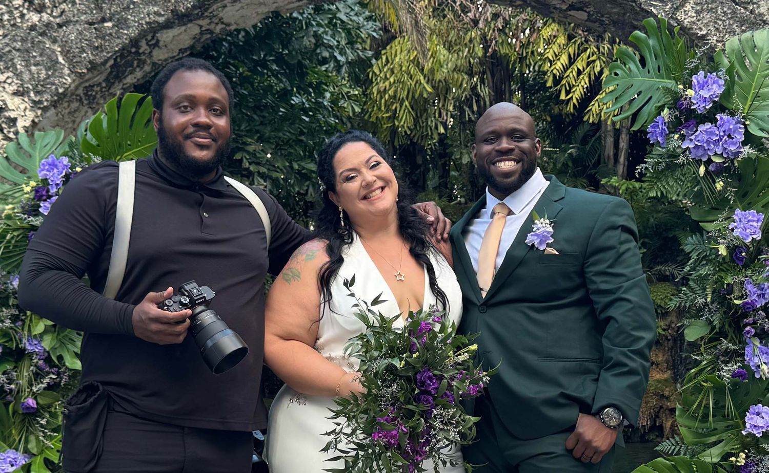 A wedding party poses outdoors in a garden with purple flowers and greenery in the background.