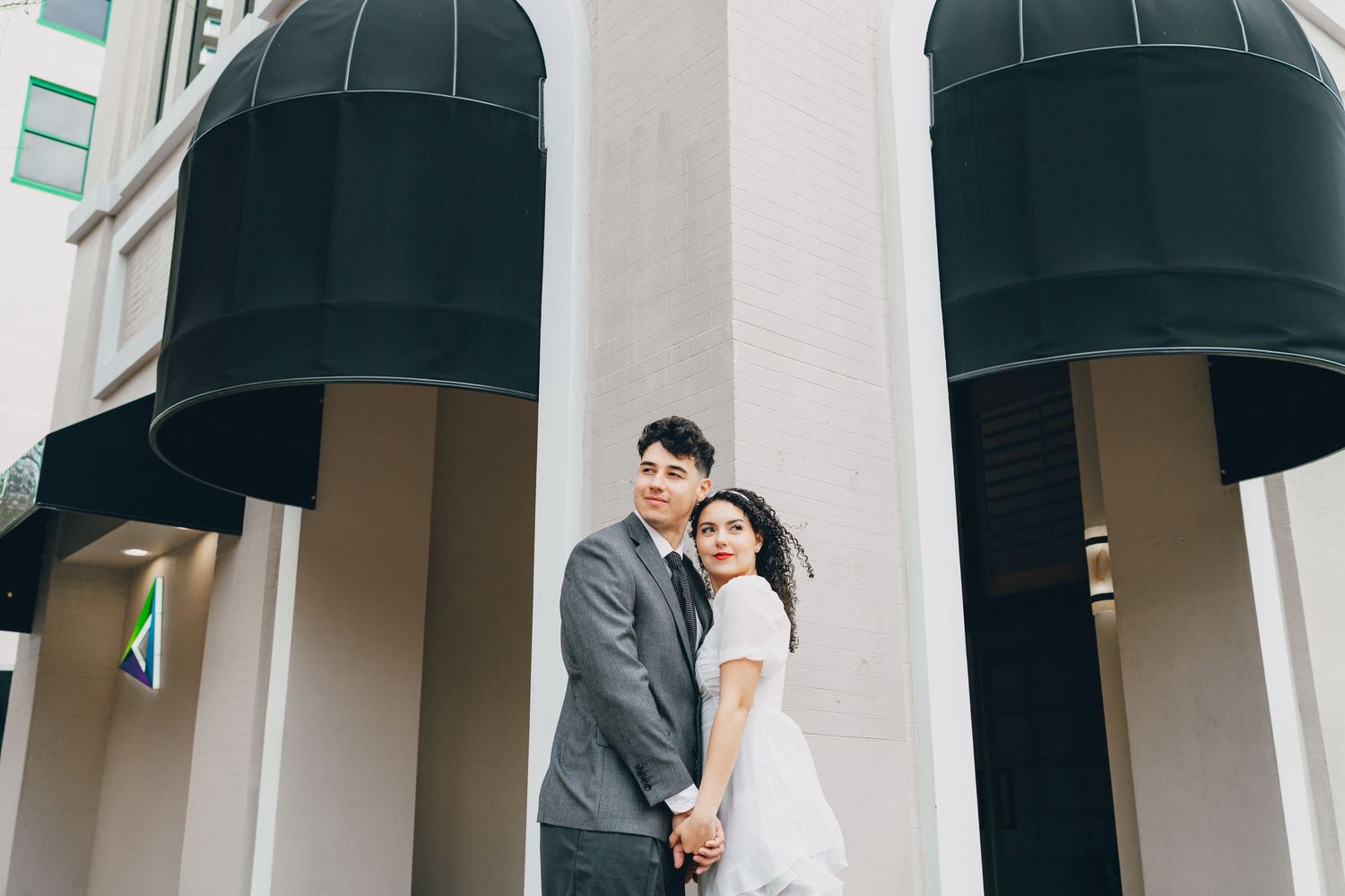 A couple stands close together under black awnings outside a modern building entrance.