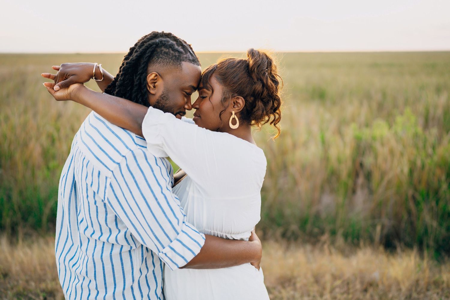 A couple shares a tender embrace in a sunlit field at golden hour wearing white and striped outfits.