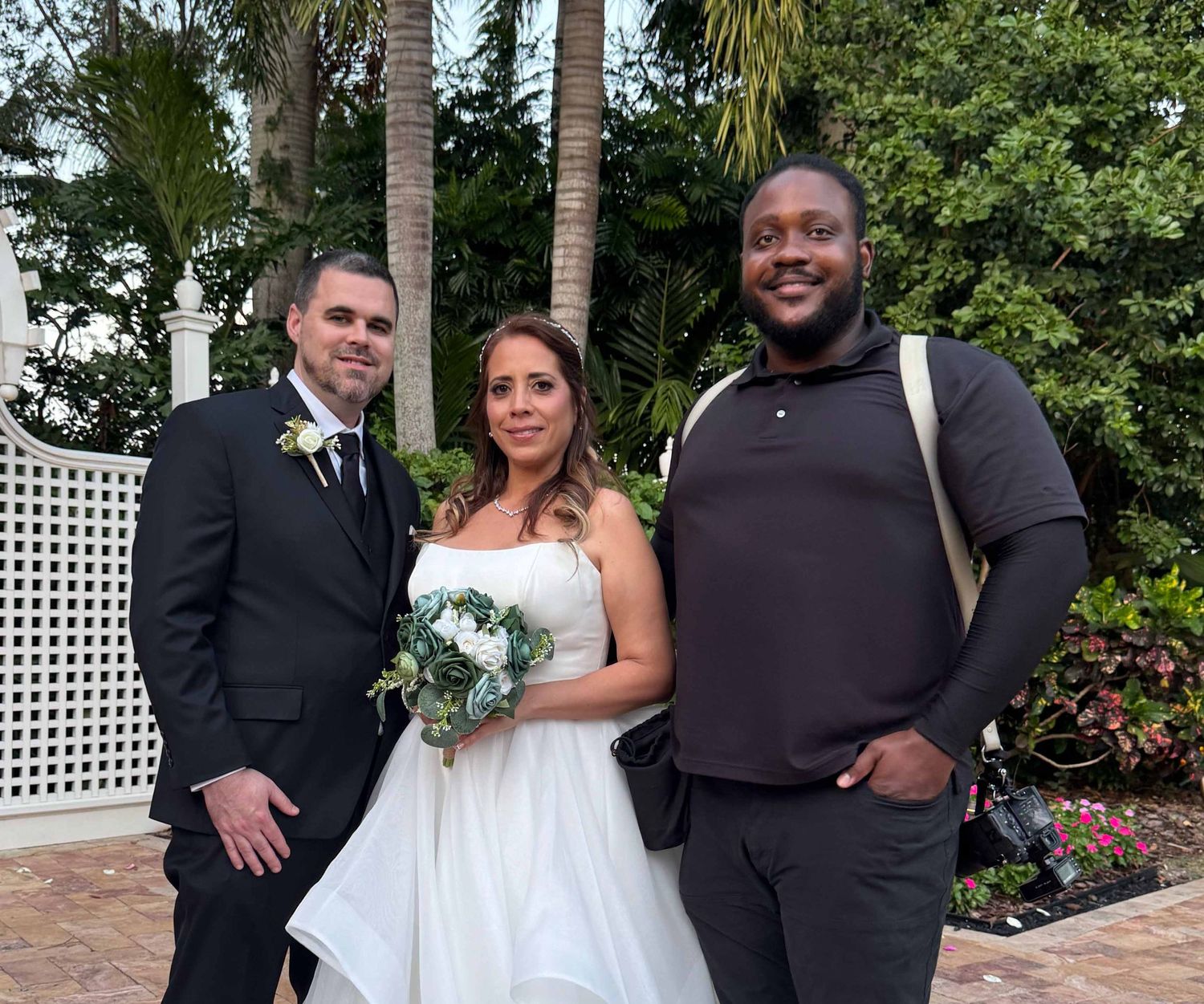 Three people pose together outdoors at a wedding ceremony with palm trees and white lattice in the background.
