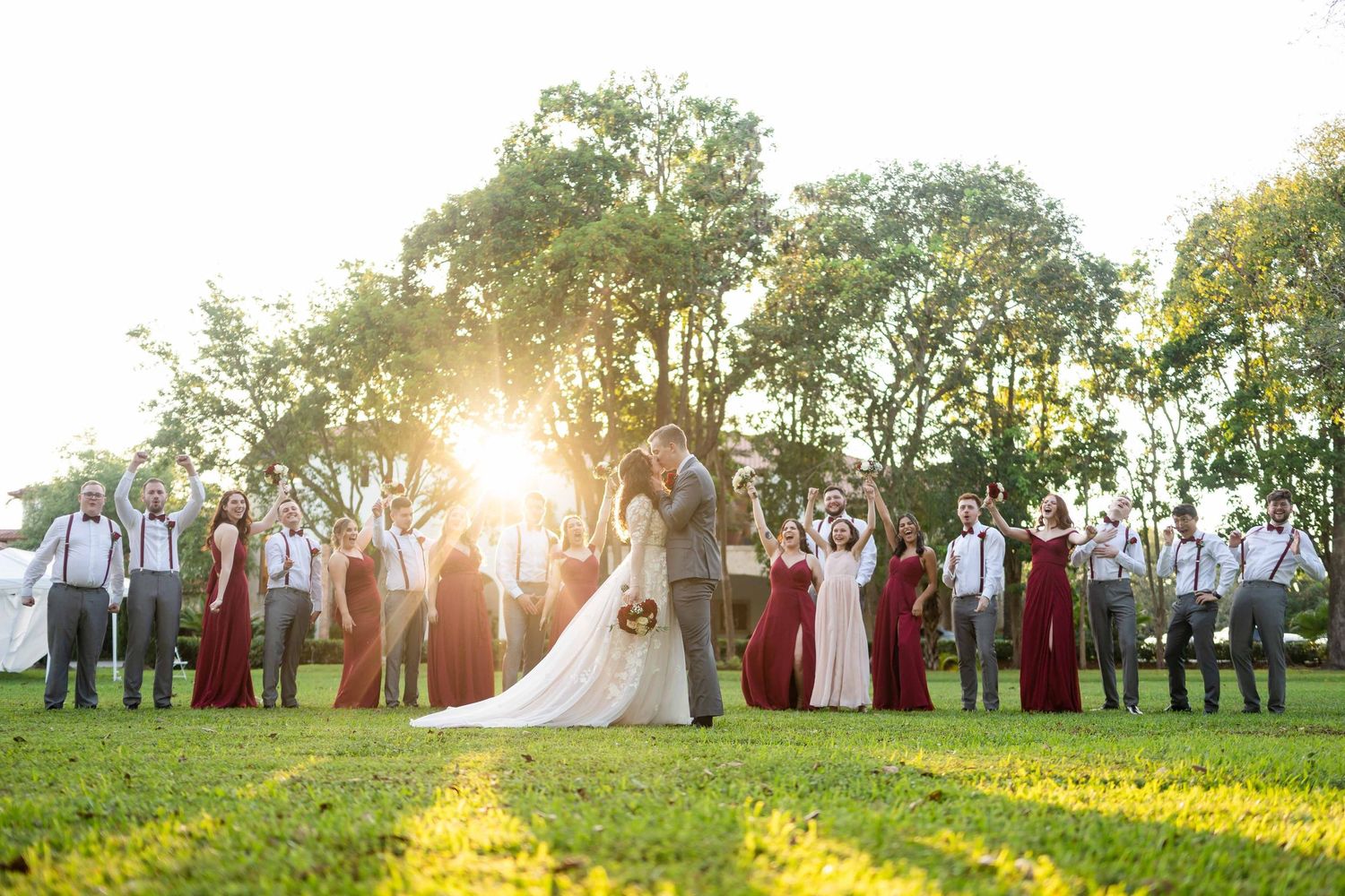 Wedding party gathers on sunny lawn during golden hour with sunburst shining through trees in background.