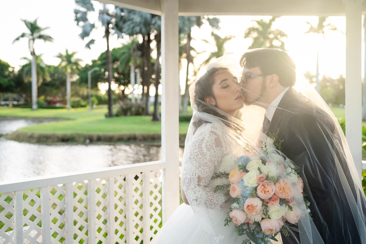 Romantic wedding couple sharing a tender moment on a gazebo near a palm-lined pond at sunset with peach floral bouquet.