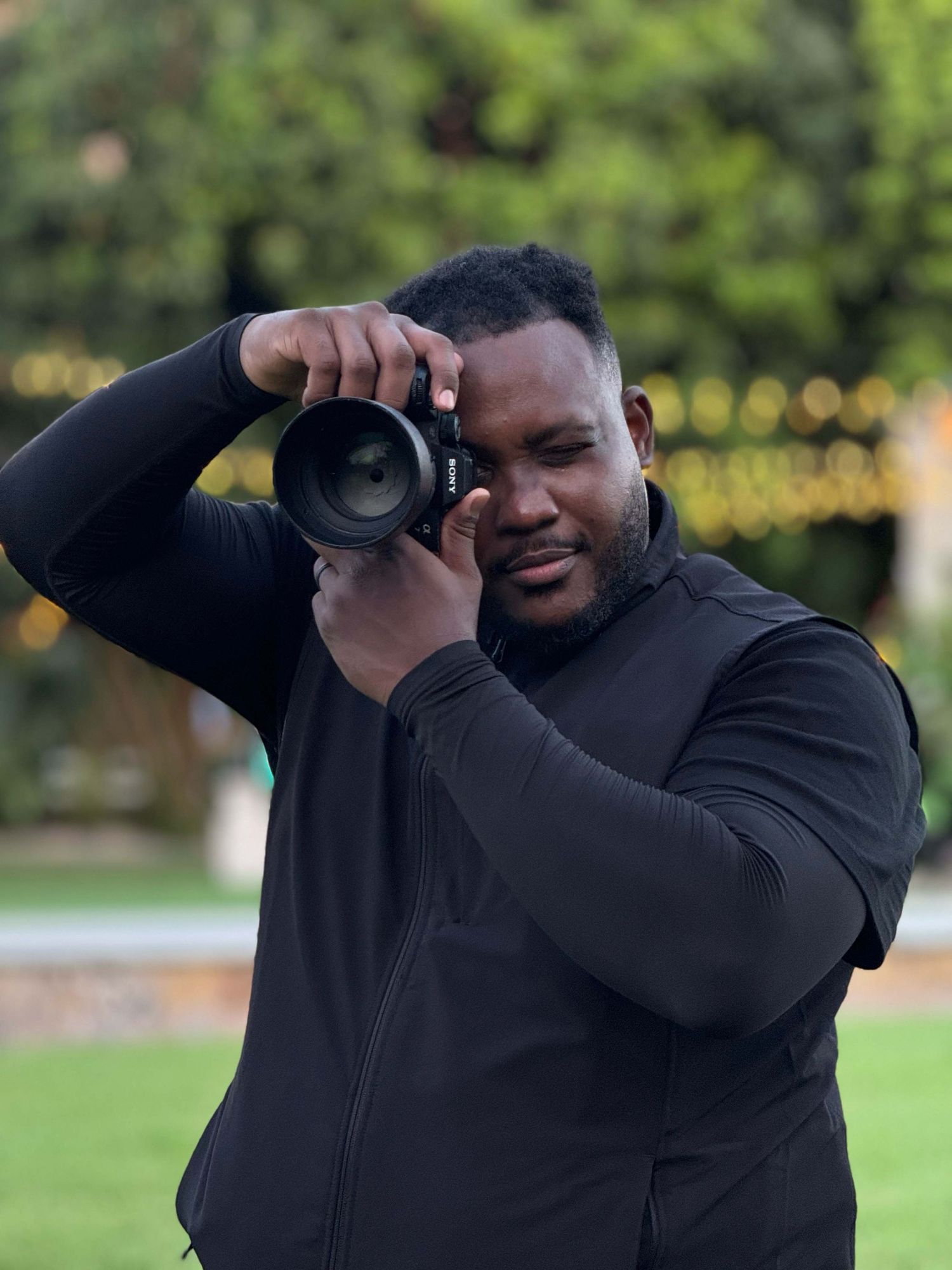 Photographer in black attire uses professional camera to take photos in outdoor garden setting.