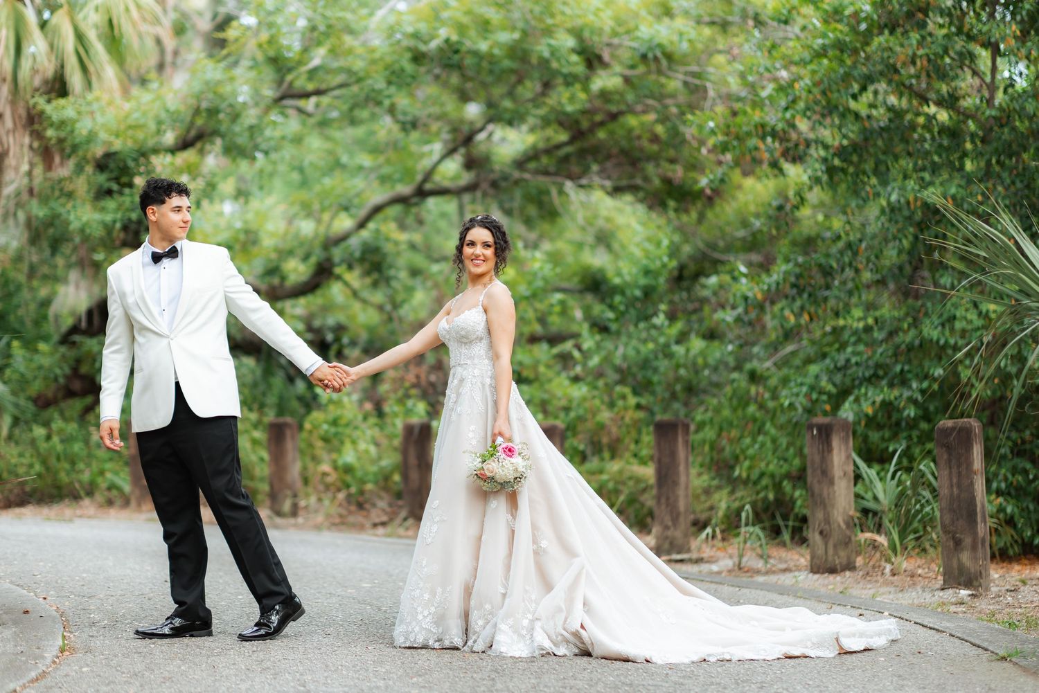 A couple in wedding attire holds hands while walking along a lush garden path surrounded by greenery.